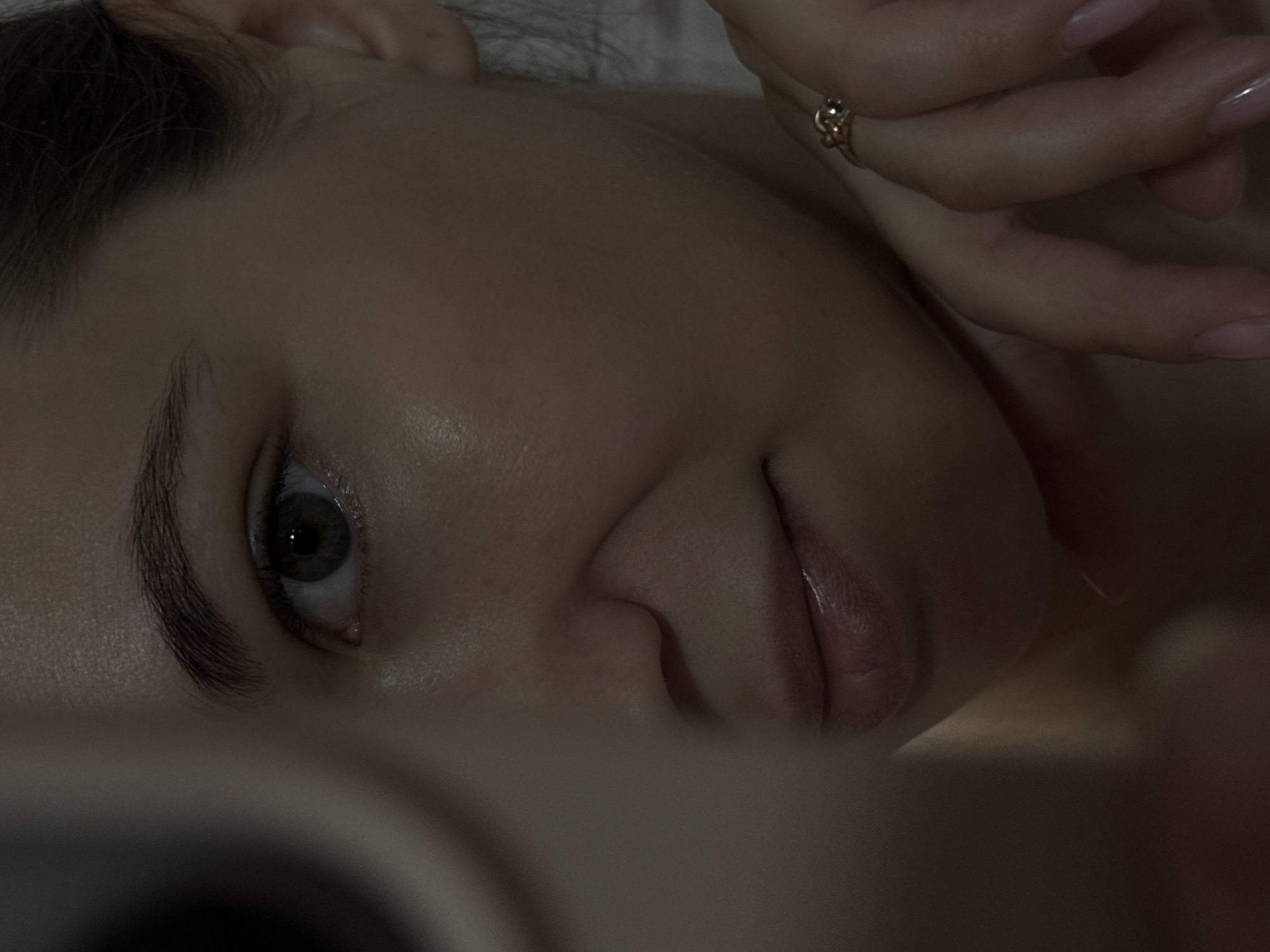 This is a close-up, waist-up, low-angle, indoor portrait of a young woman in Chaikovsky, Russia. The woman is partially obscured by a dark, out-of-focus object in the foreground, possibly a mirror or camera lens. Her right eye, with a green iris, is clearly visible and appears to be looking directly at the viewer. Her left cheek is illuminated by a strong light source, highlighting its smooth texture. She is wearing a gold ring with a small stone on her right hand, which is resting on her chin. Her fingernails are long and painted with a light pink polish. The background is dark and indistinct, with some subtle vertical lines that might suggest a wall or curtain. The overall mood of the image is intimate and enigmatic.
