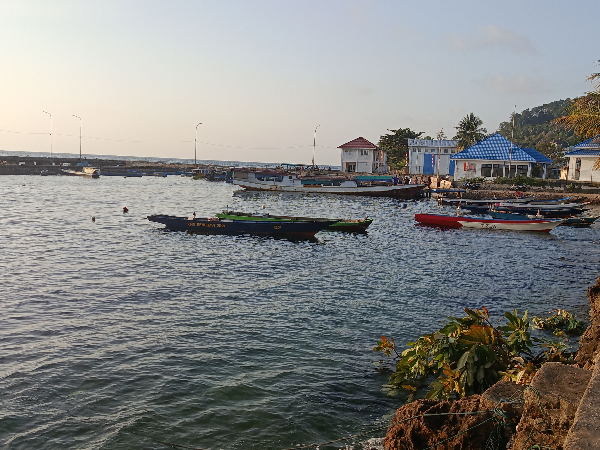 The image depicts a harbor scene in Mansalean, Indonesia, characterized by calm waters and a clear, pale sky. Several boats are anchored in the water, ranging in size and color. In the foreground, a dark green boat with white lettering and the number "02" is visible. Further back, a red and white boat labeled "TZEA" is also present.
Along the shoreline, buildings with red and blue roofs suggest a coastal community or facility. A long pier with lampposts extends into the water, with more boats moored along it. The environment appears tranquil, with the sun casting a gentle light on the scene. The presence of boats and buildings indicates that this is likely a working harbor, serving the local population and possibly for transport or fishing activities. The vegetation on the right side of the image, including some leafy branches, suggests a tropical or subtropical climate.