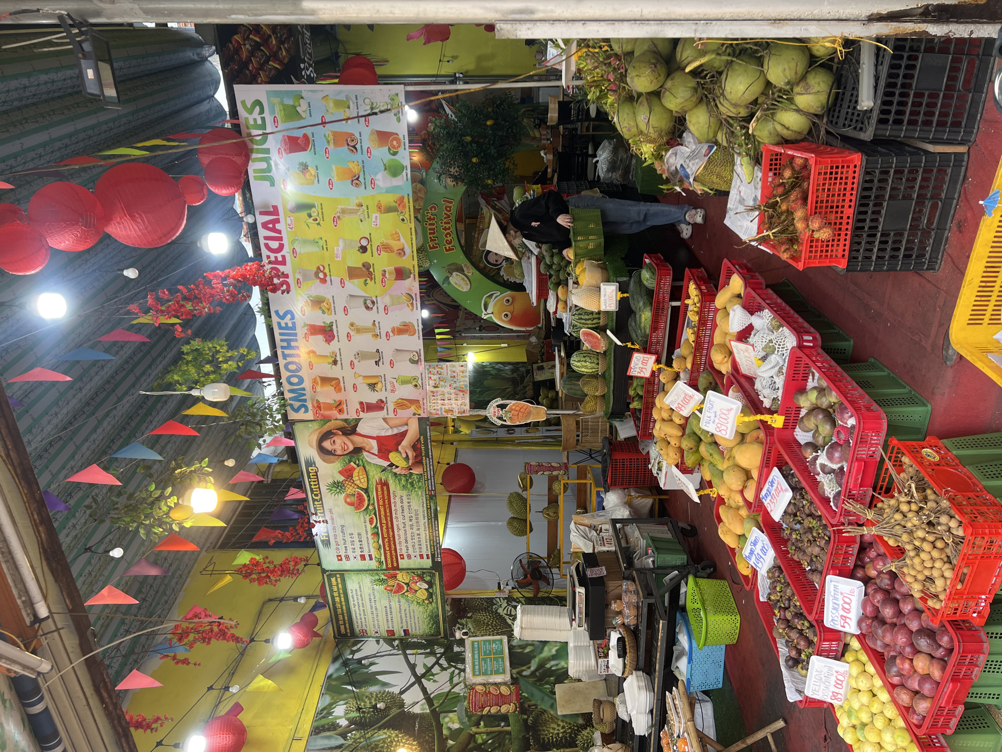 Here's a detailed description of the image:
**Scene Overview:** The image depicts a vibrant fruit stand in what appears to be Nha Trang, Vietnam. It is filled with a variety of fresh fruits, arranged for sale. The setup includes signage advertising smoothies, juices, and free fruit cutting.
**People:** A person in a black jacket is partially visible near the back, likely a customer or vendor. A woman's picture is used on a poster.
**Subjects & Objects:**
* **Fruits:** Abundant displays of mangoes, watermelons, coconuts, rambutan, longan, mangosteen, passion fruit, dragon fruit, and other tropical fruits are visible.
* **Signage:** Posters with images of smoothies and juices, along with descriptions and prices. Also promotional material for fruit cutting and claims about fruit quality.
* **Containers:** Red and green plastic crates/baskets are used to display the fruits.
* **Decorations:** Vietnamese-style lanterns, colorful flags, and artificial floral decorations are present.
* **Other:** A fan, disposable cups, containers, and other supplies for preparing smoothies and juices are visible.
**Actions & Interactions:** The focus of the scene is the display and sale of fruits. The person in the black jacket appears to be interacting with the vendor/fruit arrangement.
**Environment:**
* **Location:** Likely Nha Trang, Vietnam, based on the produce and the Vietnamese signage.
* **Setting:** A covered market area or street stall with a slightly chaotic, busy atmosphere.
* **Lighting:** A mix of natural and artificial lighting, giving the stall a warm and inviting glow.
**Forensic Details:**
* Prices are listed in VND (Vietnamese Dong).
* The fruit appears fresh and well-arranged.
* Multiple signs are present, advertising products and services in English, Korean, and Vietnamese.
* Some fruits have tags displaying their names and prices.