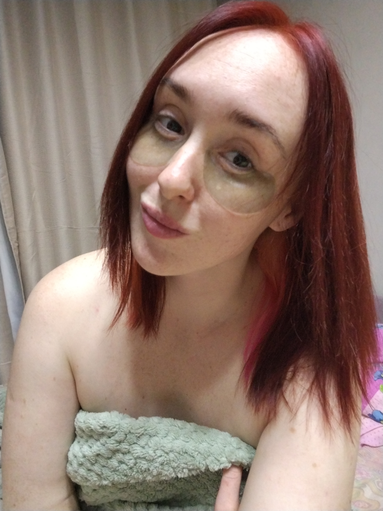 A woman with auburn hair and green eye masks is photographed indoors. She is partially covered with a green towel, appearing to be topless. The background consists of a neutral-colored curtain. The woman is looking at the camera with a slight smile. There is no indication of this photo being in Izhevsk, Russia.
