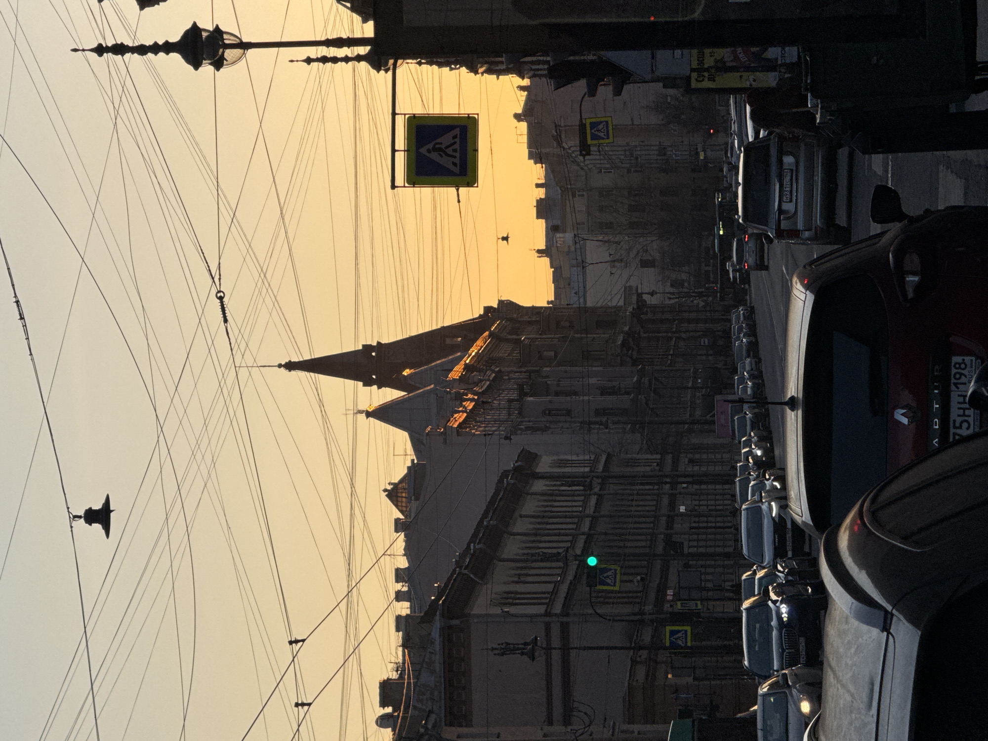 A street in Saint Petersburg, Russia, shows buildings with spire-like structures against a golden sunset or sunrise sky. Overhead power lines crisscross the sky. There is a pedestrian crossing sign visible. The street is lined with cars parked along the side, with one car having a license plate "75HH198". A traffic light is illuminated with a green light.