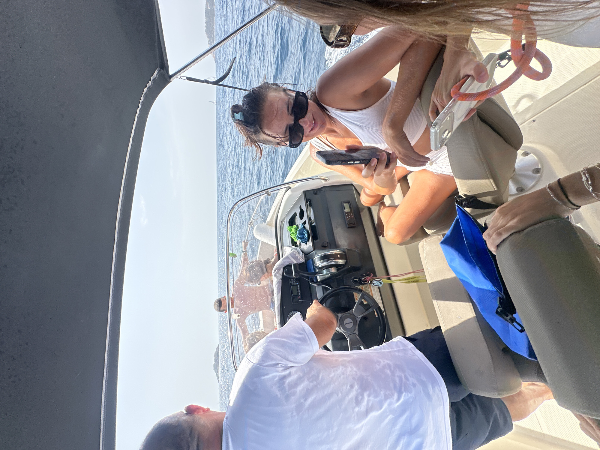 The image captures a group of people on a boat, likely a motorboat, navigating the waters of the Adriatic Sea near Luštica, Montenegro. The perspective is from within the boat, looking forward. A man in a white t-shirt and dark shorts is at the helm, his hands on the steering wheel. In the background, another man is standing, shirtless, with his arms outstretched, enjoying the ride. A woman in a white bikini and sunglasses is seated and holding a phone, seemingly taking a selfie or a photo. Another woman's arm and shoulder are visible in the foreground, also holding a phone. The water is blue and calm, with a small island visible in the distance, characteristic of the Luštica peninsula's coastline. The sky is bright and clear, indicating a sunny day. Essential elements include the boat's interior with seats and dashboard, the sea, the sky, the people engaged in typical leisure activities on a boat, and the scenic natural environment.