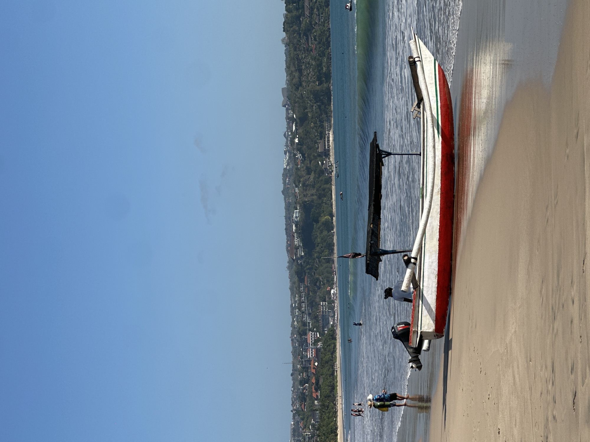 A wide shot captures a serene Kuta beach scene in Bali, Indonesia, under a clear blue sky. In the foreground, a traditional Indonesian jukung fishing boat, painted white with a red hull and green trim, rests on the wet sand at the water's edge, its outboard motor prominent. Two figures are near the boat: one in a white shirt, facing away from the camera, and another, seen from the back, wearing a dark head covering. Further down the beach, two individuals, one in a yellow shirt and sun hat and the other in a blue shirt and dark shorts, stand near the water. The ocean presents gentle waves, with a few scattered individuals wading and swimming in the distance. The background reveals the vibrant Kuta coastline, dotted with buildings and lush greenery, characteristic of the popular tourist destination.