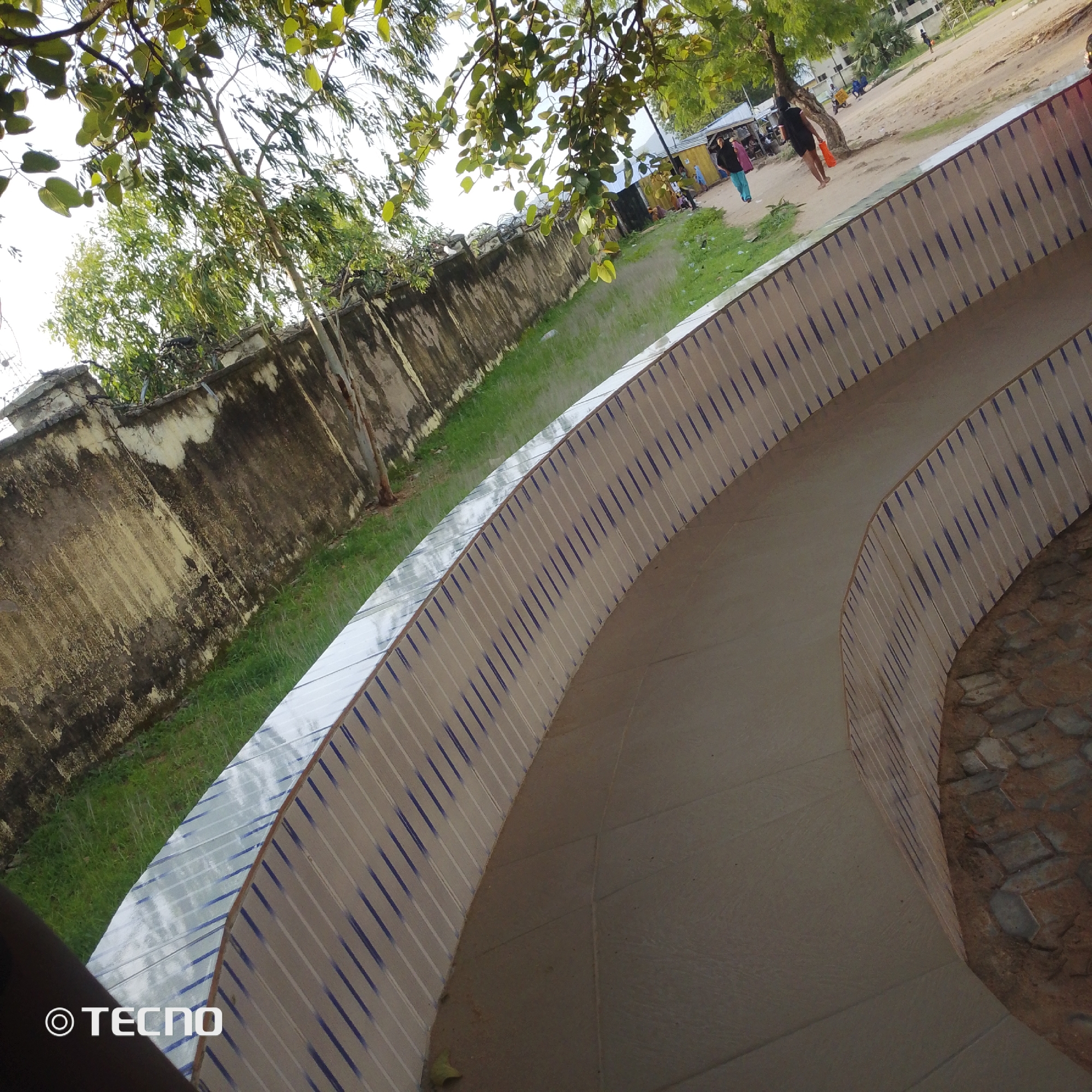 The image depicts an outdoor scene, likely in Jalingo, Nigeria. In the foreground, a tiled, curved walkway with blue and white stripes dominates the lower portion of the frame. To the left, a weathered, rough-textured wall runs diagonally upwards, with some greenery and tree branches visible above it. Beyond the wall, a grassy area is visible, leading to a dirt path or road. Further in the background, several structures, including what appear to be small shops or market stalls, are present. People are seen walking or standing near these structures, and some vehicles, including a blue tricycle-like vehicle and a motorcycle, are parked or in motion. The overall impression is of a bustling, perhaps somewhat informal, urban environment with elements of nature interspersed. A logo in the bottom left corner reads "© TECNO."