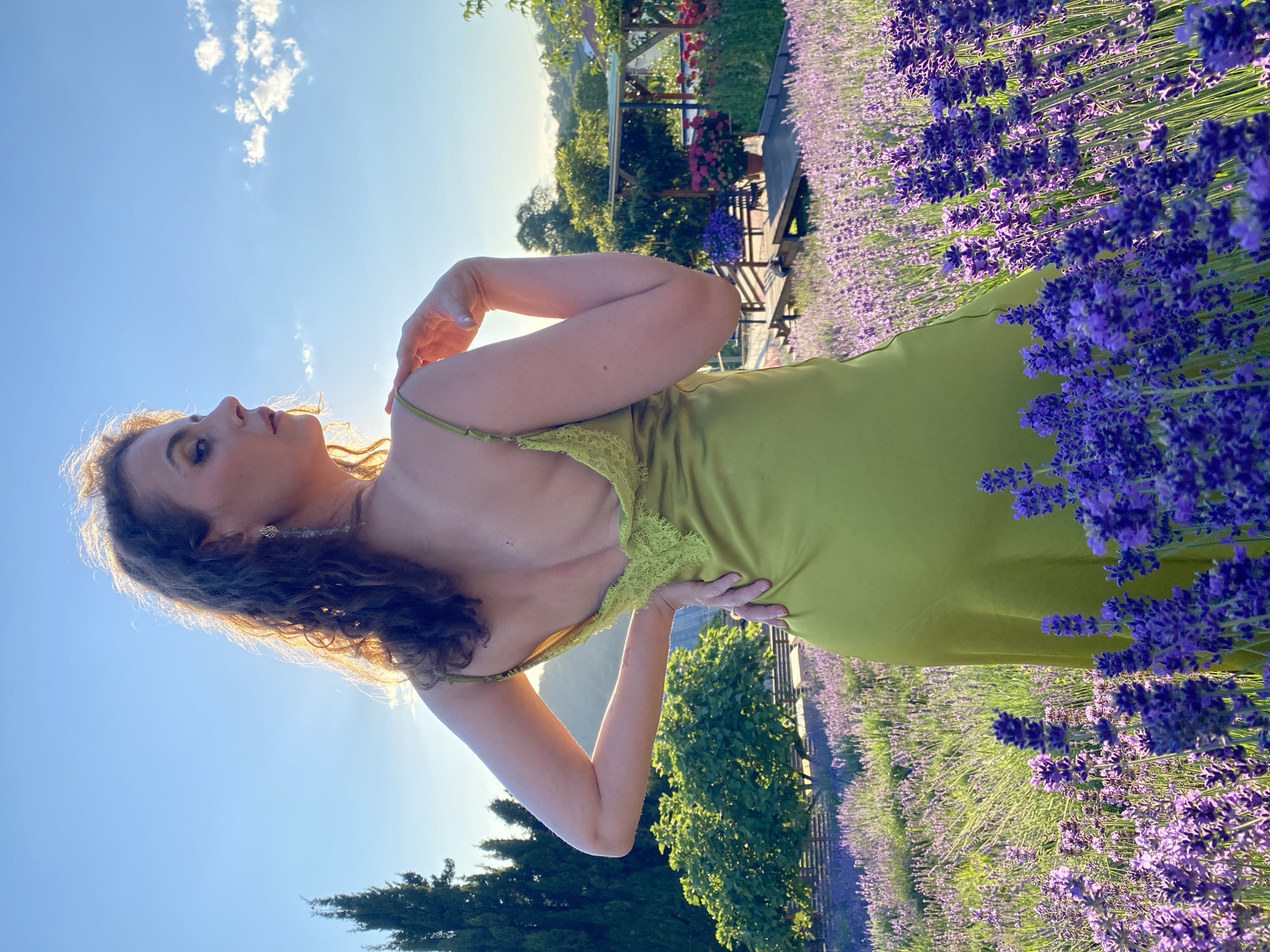 A young woman with curly, blonde hair and light makeup stands in a field of purple lavender. She is wearing a lime-green slip dress with lace detailing on the bust. Her left arm is bent, with her hand resting on her shoulder, and her right hand is on her hip. She is looking up and to her left, with a serene expression.
The sun is low in the sky, casting a warm, golden light on the scene, suggesting it is either sunrise or sunset. The sky is a clear blue with a few wispy clouds. In the background, there are trees, a wooden structure with red flowers, and more lavender fields stretching into the distance. The overall mood is peaceful and idyllic. The location is Verkhneye Uchdere, Russia. There is no visible text in the image.