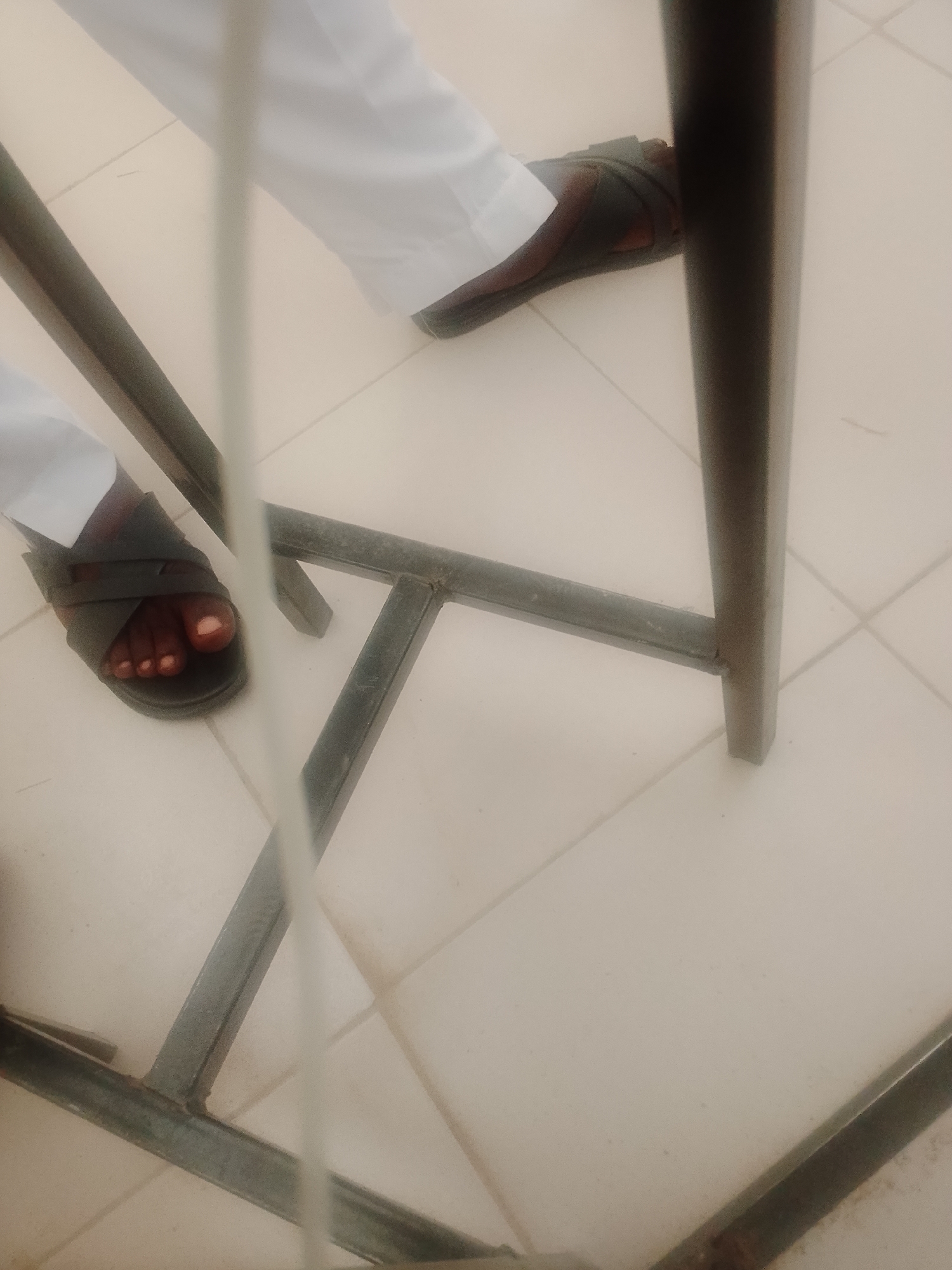 The image depicts a close-up, angled view of a person's feet wearing dark sandals, standing on a tiled floor. The person is also wearing white trousers or a robe. The feet are positioned near a dark metal structure, possibly part of a table or chair, which runs diagonally across the frame. The lighting is somewhat diffused, and the overall scene has a slightly blurry quality. The context of this image, including its location in the city of Unknown, Unknown, cannot be determined from the visual information provided.