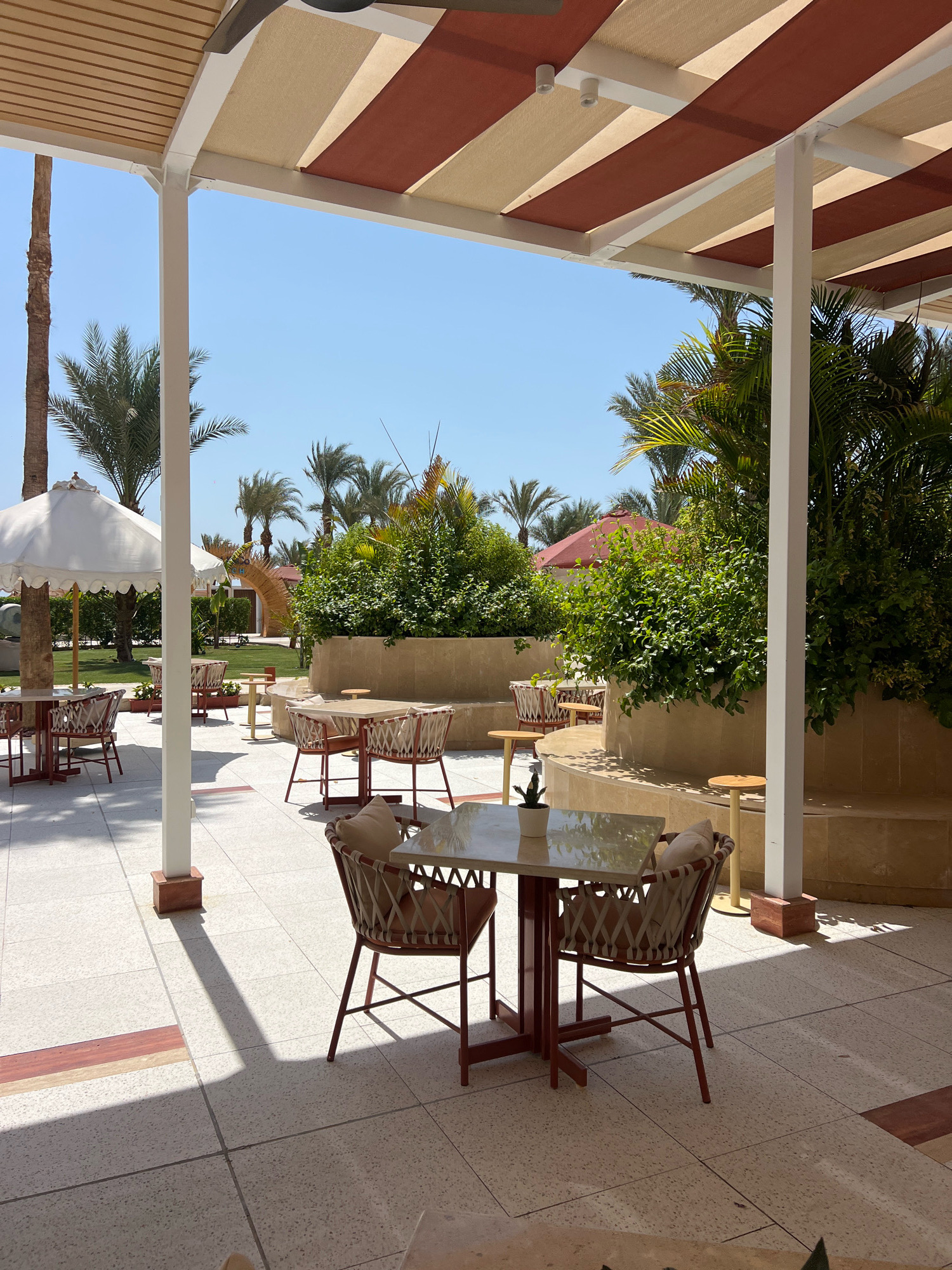 This image depicts an outdoor seating area with tables and chairs, situated on a tiled patio. The area is covered by a canopy with alternating stripes of tan and reddish-brown fabric. Palm trees and lush green bushes are visible in the background, suggesting a warm, possibly tropical or sub-tropical climate. In the distance, a red umbrella and a decorative archway can be seen. The scene is illuminated by bright sunlight, casting distinct shadows on the patio floor. The furniture consists of square tables with light-colored tops and reddish-brown metal frames, accompanied by chairs with woven seats and backs, and light-colored cushions. The overall atmosphere is that of a well-maintained outdoor dining or relaxation space, likely part of a resort or hotel in Sharm El Sheikh, Egypt, given the presence of palm trees and the characteristic sunny, clear blue sky. There are no people visible in the image.