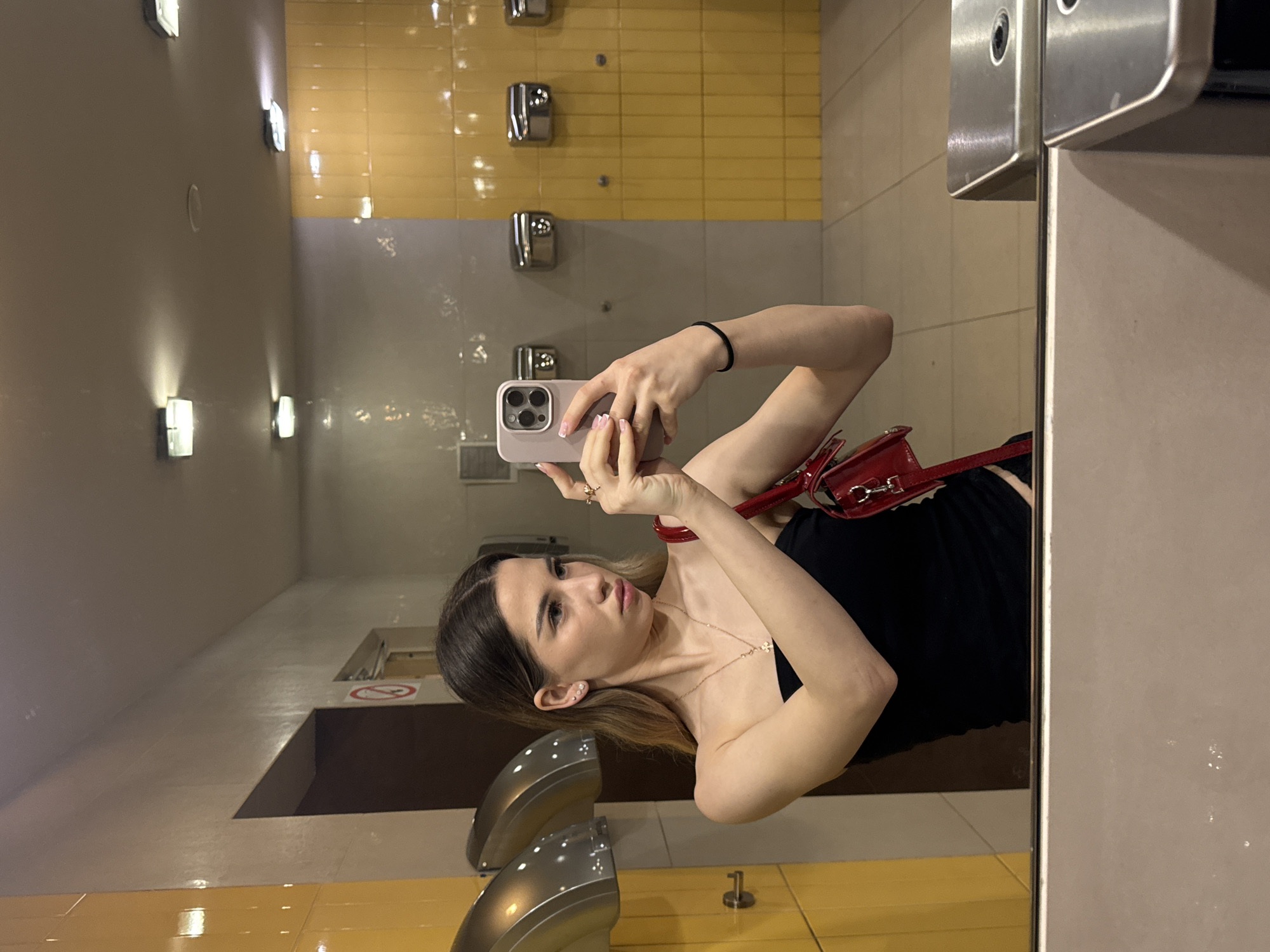 A young woman is captured in a mirror selfie, holding a light-colored iPhone in a pink/purple case. She has dark hair with lighter highlights, a black top, a gold necklace, and a red shoulder bag. A black hair tie is on her left wrist.
The setting appears to be a public restroom, identifiable by the presence of multiple chrome hand dryers and faucet units reflected in the mirror. The walls are tiled, featuring a band of bright yellow rectangular tiles above a section of gray/beige tiles. Several rectangular wall lights illuminate the space.
The woman is looking towards the phone, seemingly posing for the picture with a neutral expression. Part of a toilet stall partition is visible to the right. On a surface reflected behind her, a red circular sign with a diagonal line through it, commonly indicating a prohibition, can be faintly seen. The flooring is also tiled, matching the lower wall color in some areas. The overall lighting is artificial. The image is presented sideways.