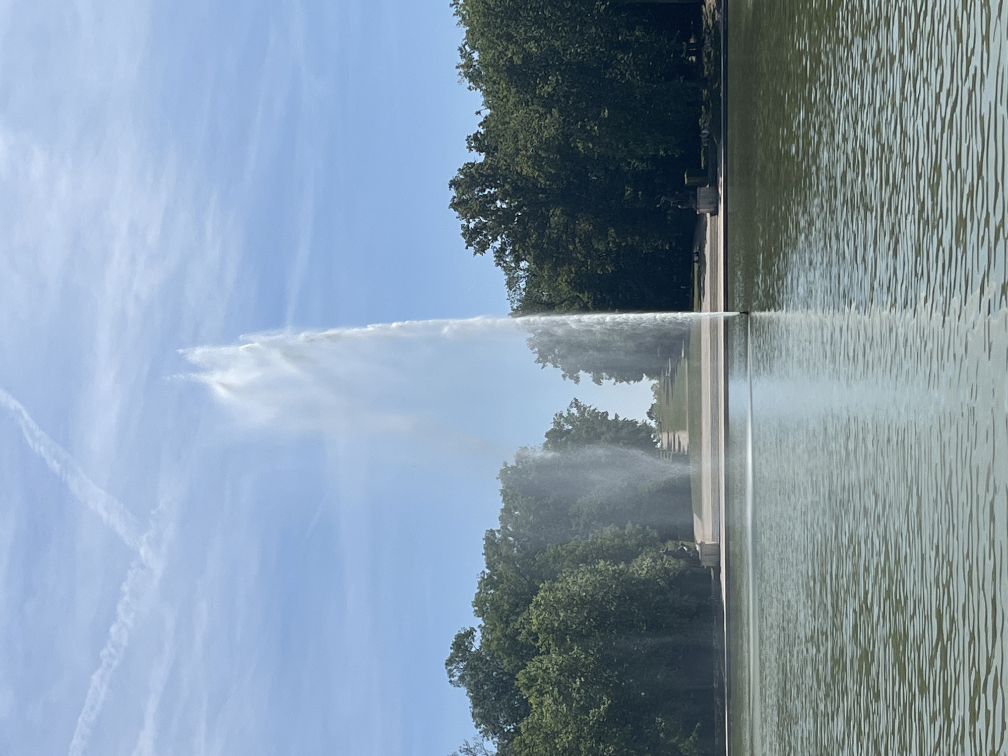 The image depicts a large fountain shooting a powerful stream of water into the air, with a reflective pool in the foreground. The fountain is situated in a park-like setting with lush green trees lining the background. The sky is a clear blue with a few wispy clouds. In the distance, there appears to be a series of steps leading up a gentle slope, possibly part of the park's landscaping. There are also some dark figures, likely statues or people, visible on the banks of the pool. The scene suggests a public space, likely a park or garden, designed for recreation and enjoyment of natural beauty.
This is the Parc de Sceaux, located in Antony, France.