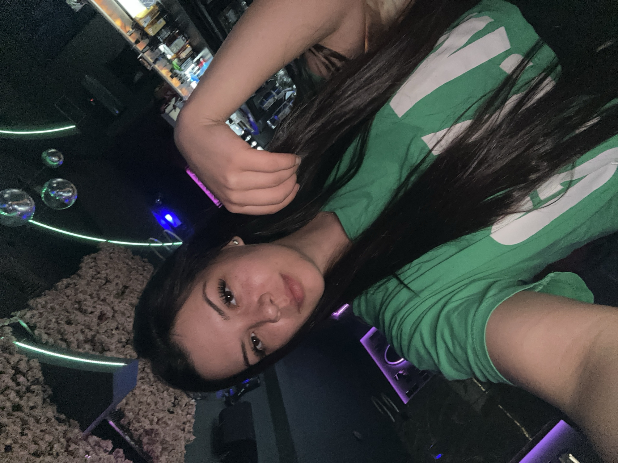 This is a selfie of a young woman with long, dark hair and light skin, wearing a green t-shirt with white lettering. She is holding her hair with her right hand. She is in what appears to be a nightclub or bar. In the background, there are disco balls hanging from the ceiling, neon lights, and a bar with bottles. The lighting is dim and atmospheric, consistent with a nightlife setting. The image does not provide any specific information about the city of Sliven, Bulgaria.