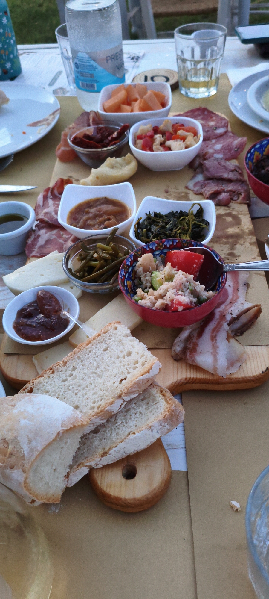 The image shows a close-up of a table laden with a charcuterie board and various accompaniments, likely set outdoors. The scene is composed of several small white bowls and a decorative red bowl filled with different food items, including sliced meats, cheese, olives, green beans, a salad-like mixture, and what appears to be jam or a relish. Slices of rustic bread are prominently featured, along with some cured meat and bacon. A bottle of water, a glass of white wine, and a small plate are also visible. The table surface is a light-colored wood. In the background, a green and white patterned bottle and some indistinct outdoor elements suggest a natural setting. The overall impression is a relaxed, rustic meal. There is no indication of the city of Buriano, Italy, in the image.