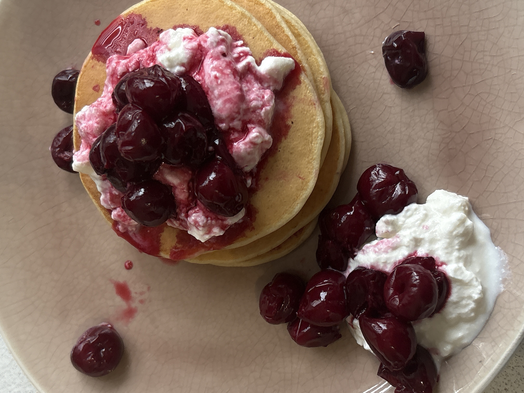 The image shows a plate of food. The plate is a light pink color with a textured surface. There is a stack of pancakes topped with what appears to be cottage cheese or yogurt and cherries. Several cherries are scattered around the pancakes and the yogurt. The food is the main subject of the image, and the setting is a table or countertop, likely indoors. There is no evidence to determine the location of Saint Petersburg, Russia.