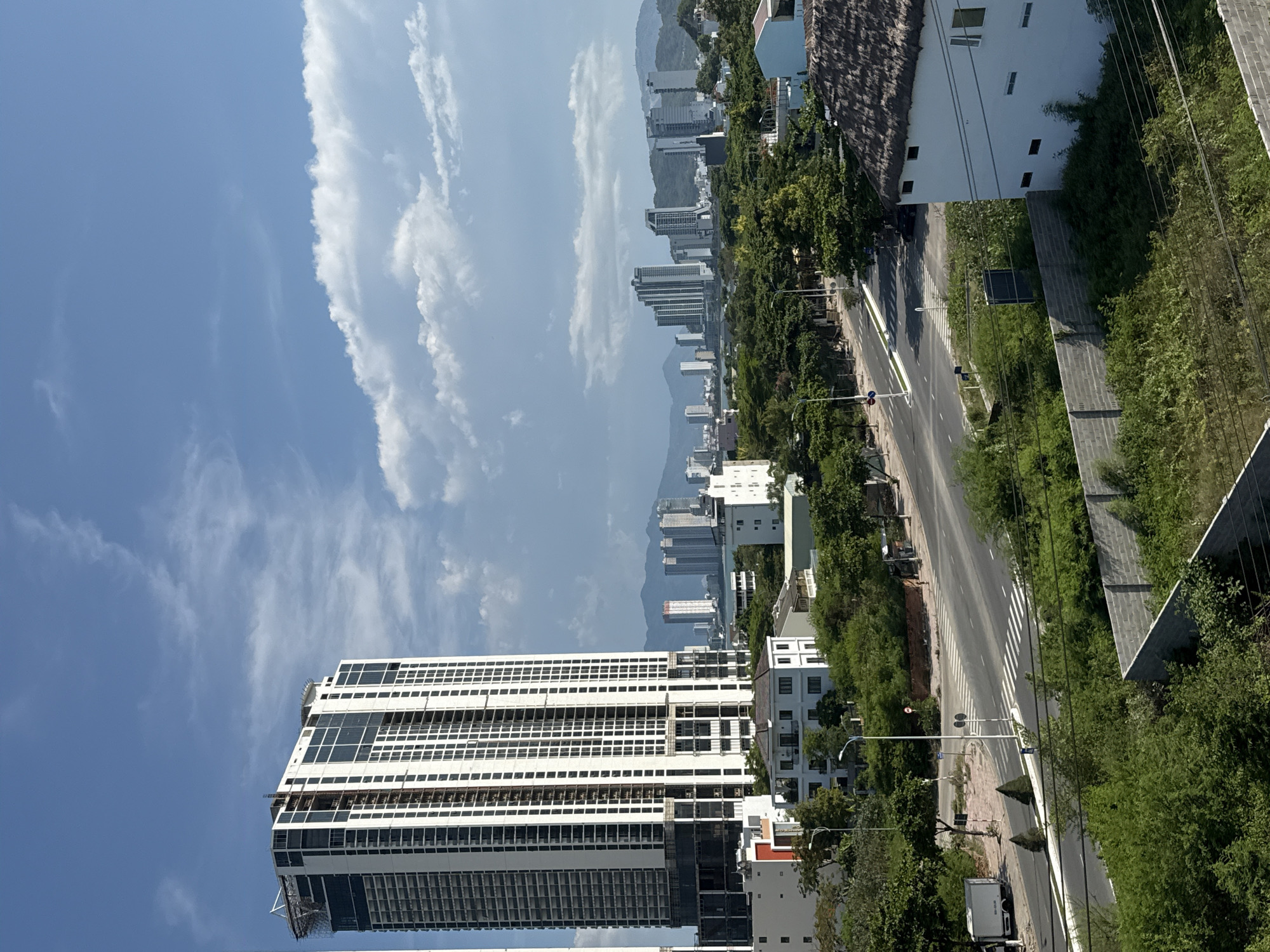 This is an outdoor, eye-level shot of a city scene in Bac Nha Trang Ward, Vietnam. The sky is a bright blue with scattered white clouds. In the foreground, there is a white building with a thatched roof and a brick wall surrounding it. To the left, a road curves through a green landscape with trees and smaller buildings. A white truck is parked on the side of the road. In the midground, a tall, modern skyscraper dominates the left side of the frame. It is a white building with many windows. Beyond the skyscraper, a cluster of taller buildings forms a city skyline. The background shows more buildings and mountains. The overall impression is one of urban development nestled within a natural landscape.