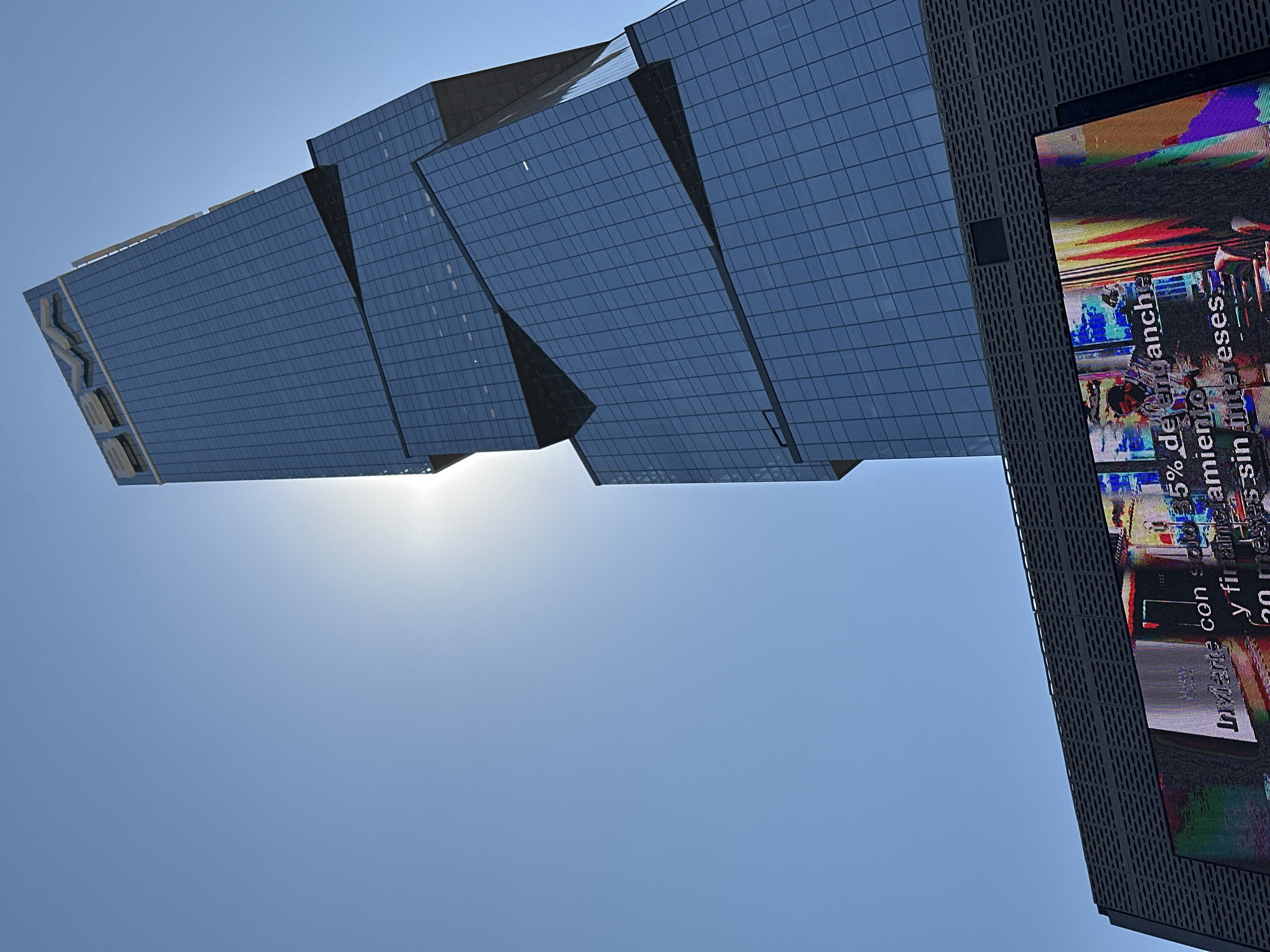 A modern, multi-tiered glass skyscraper with a segmented and offset design dominates the frame, rising against a clear blue sky. The "BBVA" logo is visible in white at the building's apex. The structure's dark blue glass facade reflects the environment, with the sun positioned behind its upper-middle section, creating a bright glare. At the base of the skyscraper, a large digital billboard displays a fragmented, vibrant advertisement with distorted imagery. Partial Spanish text on the screen includes "Invierte con...", "5% de enganche...", and "sin intereses...", suggesting a financial promotion. The billboard is integrated into a dark gray paneling with a distinct perforated pattern. No live individuals or active interactions are discernible in the physical scene. This urban setting is located in San Bernardino Tlaxcalancingo, Mexico.