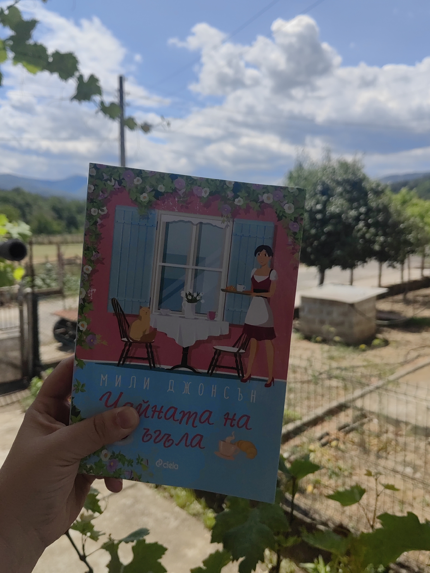 A person's hand holds a book with a brightly colored cover depicting a woman in an apron carrying a tray with pastries, a cat on a chair, and a table with flowers. The book is positioned in front of a blurred outdoor scene featuring green trees, a stone structure, and a blue sky with white clouds. The text on the book is in Cyrillic script and appears to be "МИЛИ ДЖОНС" and "ЧАЙНАТА НА ВЪЗГЛА". The background suggests a rural or semi-rural setting, possibly in the vicinity of Djerovo, Bulgaria, given the natural landscape and the style of some of the structures.