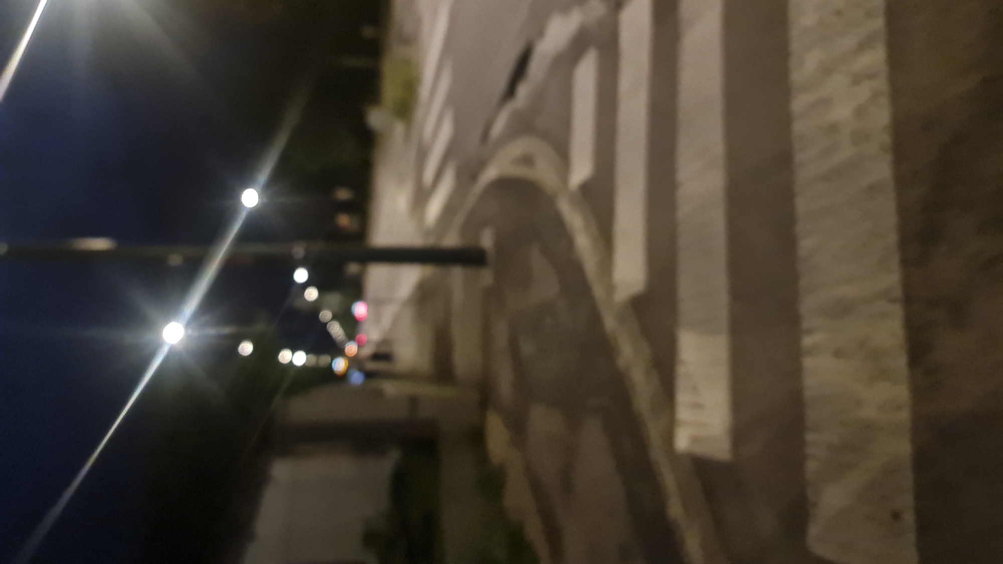 Here is a description of the image:
The image shows a blurry, low-quality street view at night. A row of blurry lights extends into the distance along one side of the street, possibly indicating streetlights or decorative lighting. Adjacent to the street is a sidewalk with several vertical, rectangular structures that are likely architectural elements, possibly pillars or retaining walls. These are made of a light-colored, textured material. The street surface has visible road markings, potentially a crosswalk or lane divisions, also in a light color. Vegetation is present along the street, partially obscured by the darkness. The sky above is a dark blue, transitioning towards black.