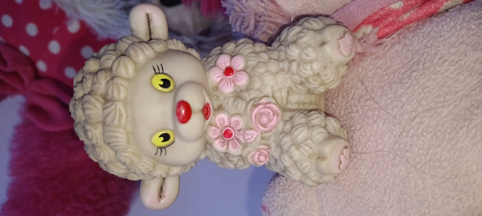 A close-up, slightly angled shot features a cream-colored rubber toy sheep with pink accents, sitting on a pink fuzzy surface. The sheep has yellow eyes with black pupils and eyelashes, a red nose, and red lips. Pink flowers with red centers are attached to its woolly body. Its feet also have pink detailing. The background is blurred and consists of various pink and fuchsia plush fabrics, some with white polka dots. The image is taken indoors, with no visible indication of the city of Rejing, Indonesia.