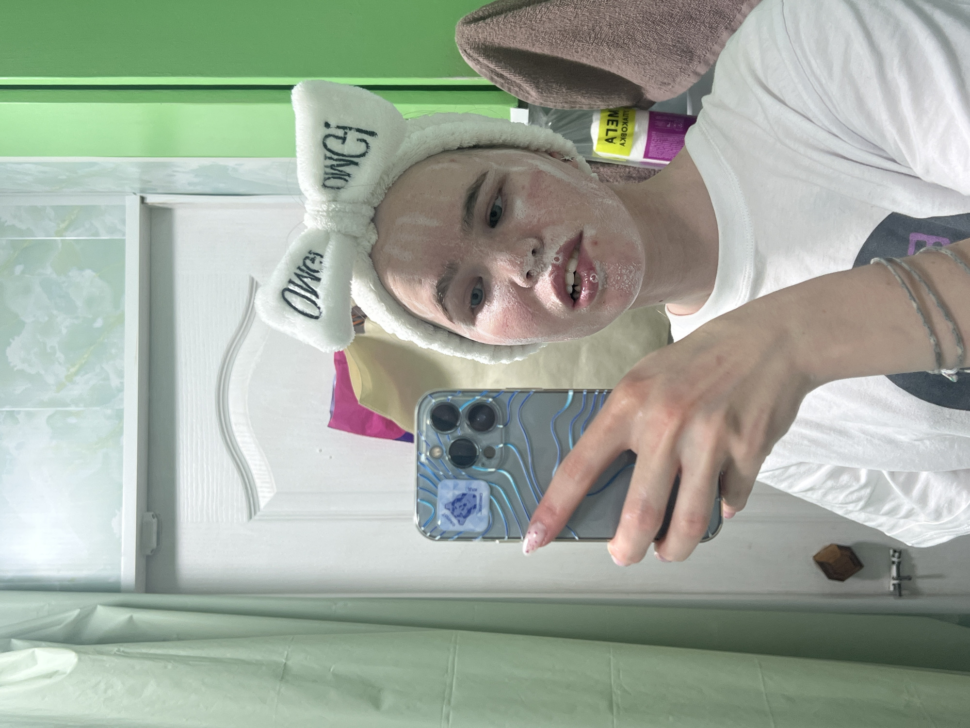 A person wearing a white bow headband with "OMG!" printed on it is taking a selfie in what appears to be a bathroom. The person's face is covered in a white substance, possibly a facial mask. They are wearing a white t-shirt. The phone has a blue patterned case and an adhesive grip on the back. The background includes a mirror, a green wall, a white door, and a shower curtain or wall covering. There's a towel and a tube of a cosmetic product partially visible. It is not possible to determine the location in the city of Chaikovsky, Russia without additional information.