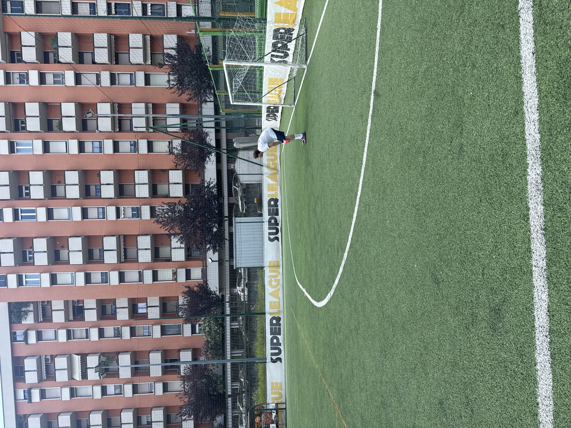 The image captures an artificial turf football pitch in an urban environment, consistent with a city like Milan, Italy, under natural daylight conditions. One male individual, likely a goalkeeper, is positioned on the right side of the frame, standing near a white-framed football goal with a net. He wears a white short-sleeved t-shirt, dark shorts, red goalkeeper gloves, and red/purple athletic shoes. He is bent forward with his hands resting on his knees, facing towards the left side of the frame.
The green artificial turf is marked with white lines, including a visible arc of the penalty area and a straight line extending from the left. In the background, a large, multi-story residential building with a reddish-orange facade and numerous white-railed balconies is prominent. Many windows are fitted with closed blinds. Dark-leafed trees and bushes are planted behind a dark metal fence that separates the pitch from a background area containing partially visible parked cars and a small grey utility structure. Along the fence line, a white banner displays the words "SUPER LEAGUE" in black and yellow lettering. A tall floodlight pole is visible on the left side of the pitch, extending above the fence. The sky above the building appears clear.