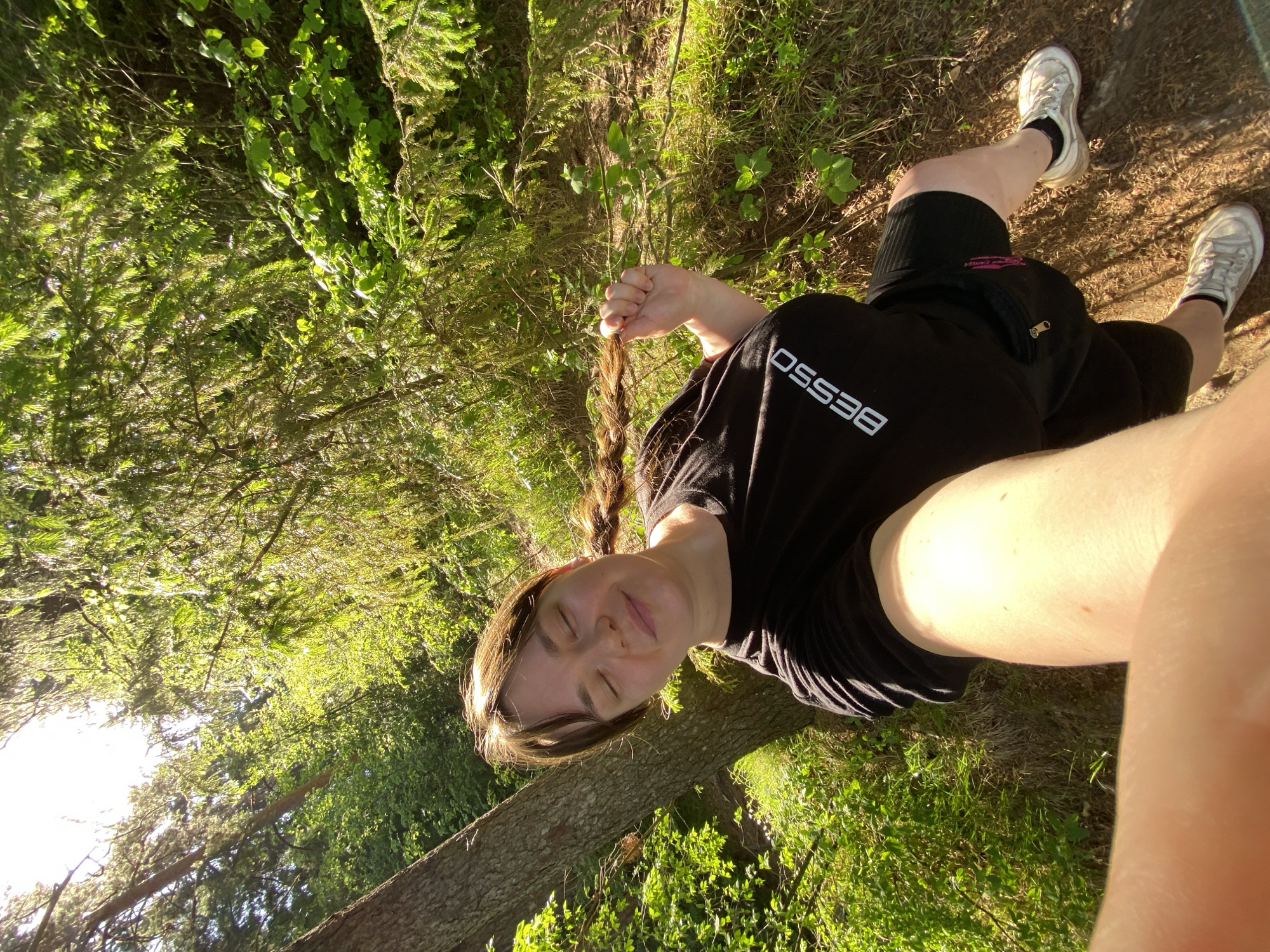The image shows a woman lying on the grass in a wooded area, likely in Votkinsk, Russia. The woman is wearing a black t-shirt with white lettering "BESSO", black shorts, and white sneakers. She has long, braided hair, and her eyes are closed. Her right hand is holding her braid. She is positioned near a fallen tree trunk. The environment consists of green grass, trees, and foliage. Sunlight is filtering through the trees, creating a dappled light effect. A fanny pack is strapped around her waist.
