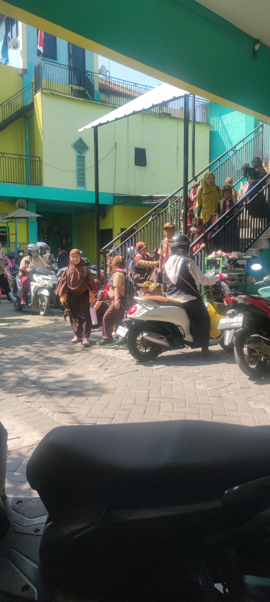 This is a vertical, full shot of a street scene in front of a building with a green and yellow facade, likely a school, in Banjarsari, Indonesia. The ground is paved with grey bricks. In the foreground, the dark seat of a parked motorcycle is visible. Several people, predominantly children wearing brown uniforms and some adults, are present. Many of the children appear to be students, some wearing scouting attire. They are gathered near a staircase leading up to the building, and some are interacting with adults. Several motorbikes are parked on the street, with one white scooter in the mid-ground with a person wearing a helmet and black pants riding it. Another red motorbike is partially visible on the right. The building has multiple levels with balconies and stairs. The lighting suggests it is daytime, with shadows cast on the brick pavement.