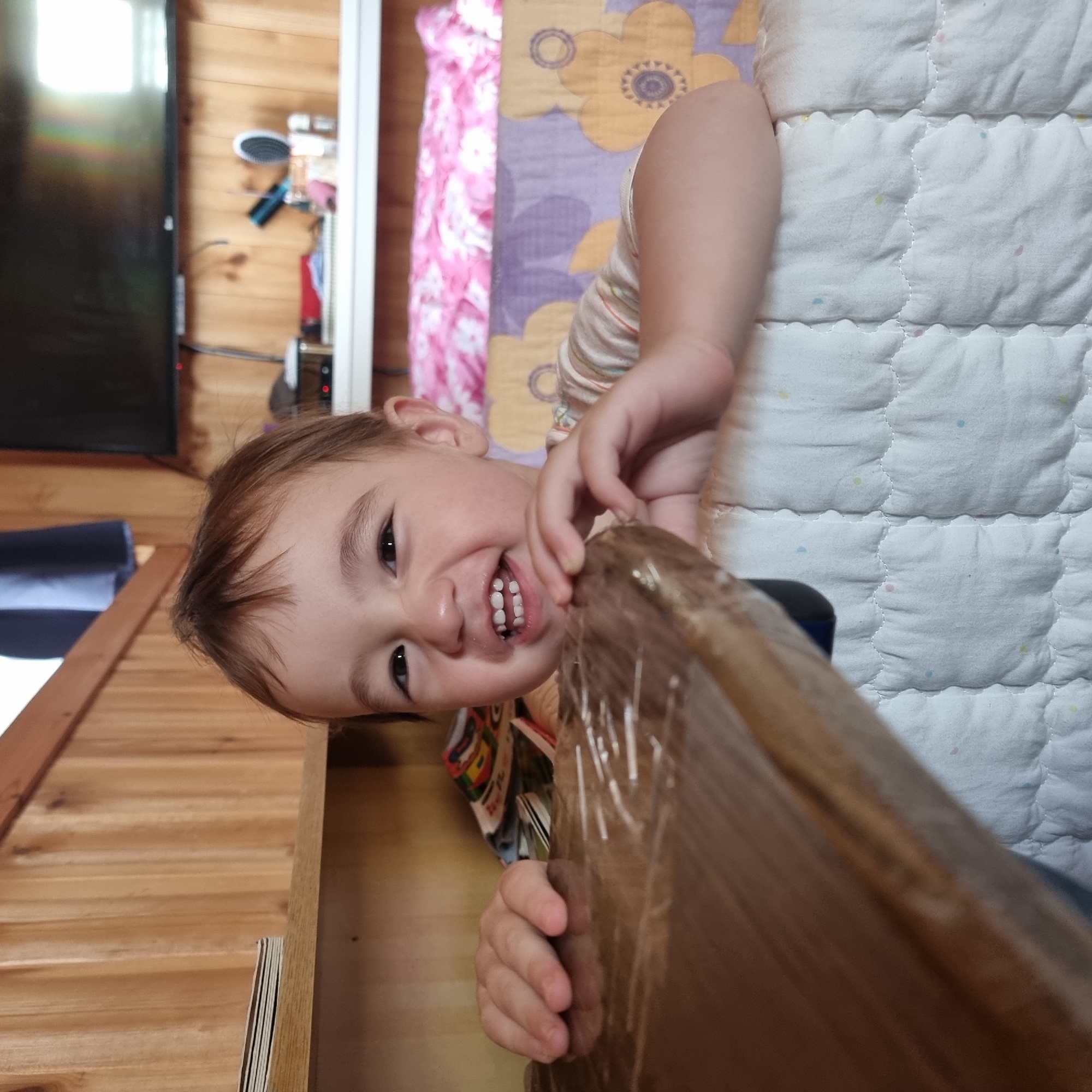 A child is peeking over a wooden surface, smiling with teeth showing. The child has brown hair and is wearing a striped shirt. The background consists of wooden walls and a shelf with some items. A television is visible on the wall above the shelf. To the right, there is a bed with a floral pattern. The scene appears to be indoors, possibly in a domestic setting. No specific location in Dunnae-myeon, South Korea, can be determined from the image.