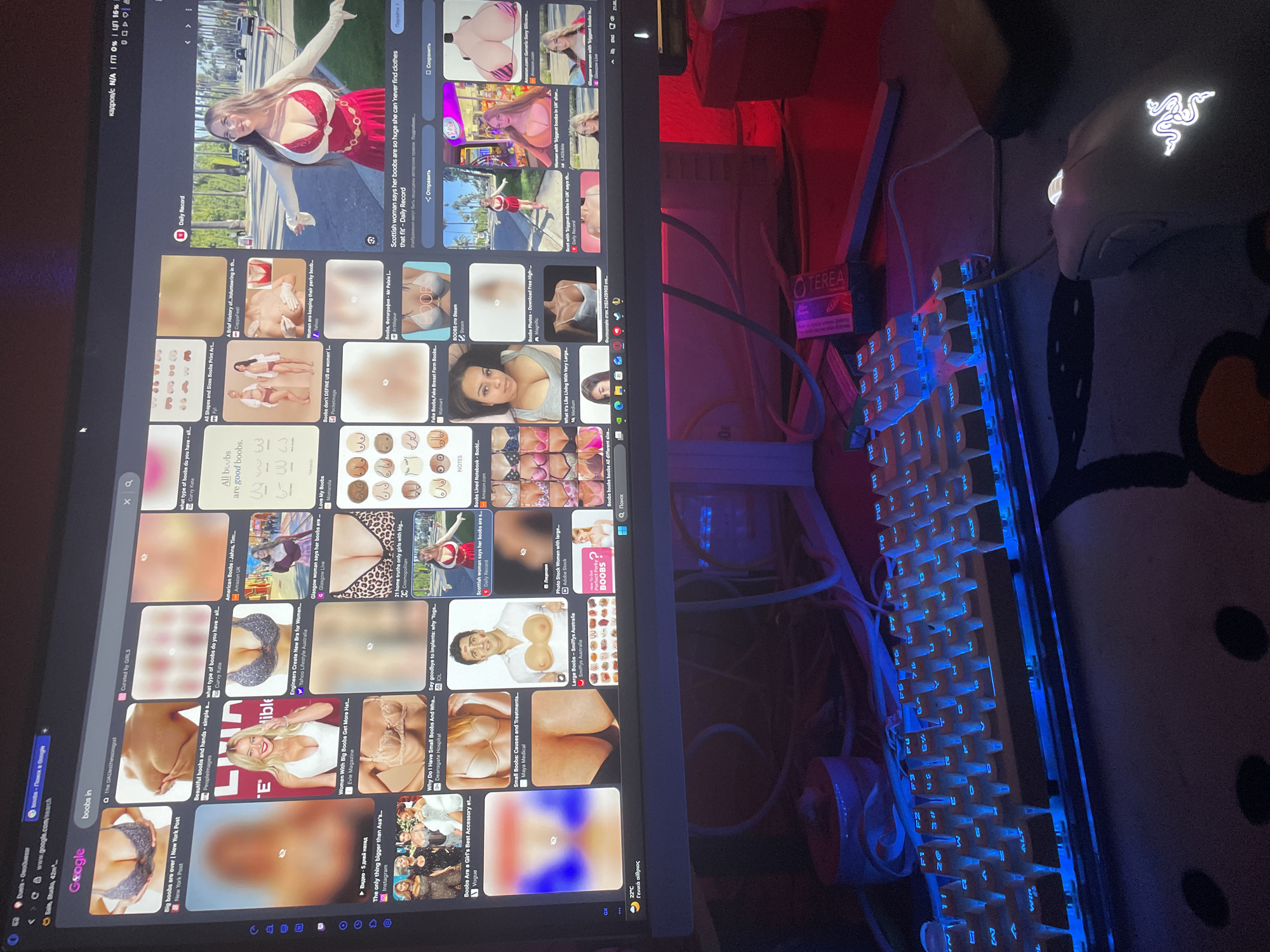 The image displays a computer monitor showing a Google search results page for "boobs in". The results include various images, articles, and links related to breasts, with some images blurred for content moderation. The monitor is positioned on a desk, with a backlit keyboard and a mouse in the foreground. The environment appears to be a home office or personal computer setup. There is no identifiable location in Neoi Epivates, Greece, in this image.