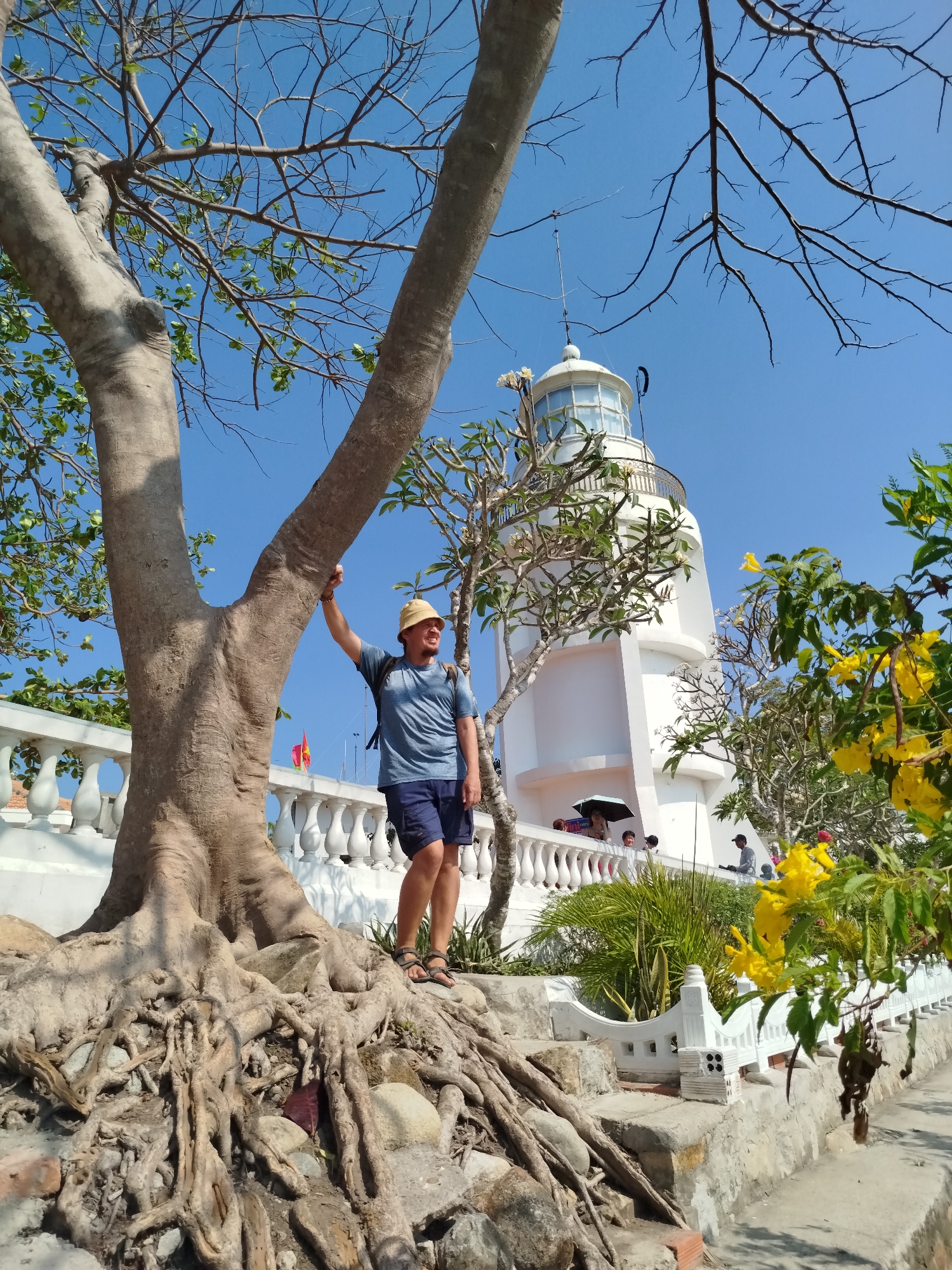 A medium shot captures a man standing next to a tree, with a lighthouse visible in the background. The man, wearing a blue t-shirt, blue shorts, sandals, and a straw hat, has a backpack and is holding onto the tree trunk. The tree has a large trunk and visible roots spread across the ground. Behind the man and tree is a white lighthouse structure. A white railing is visible along with other plants and greenery. The sky is clear and blue. The location is Ho Chi Minh City, Vietnam.