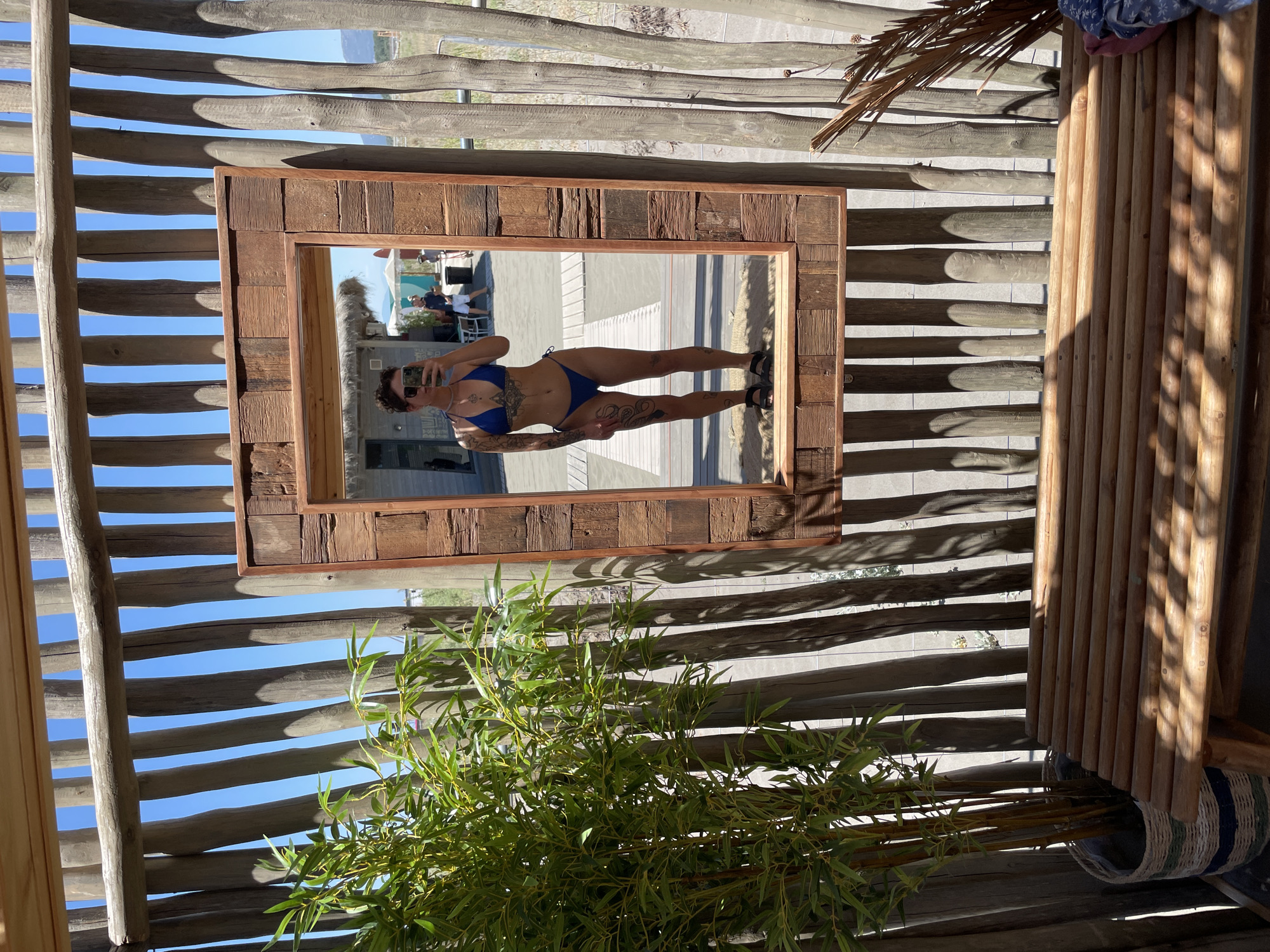 In Donji Štoj, Montenegro, a person is taking a mirror selfie in a rustic outdoor setting. The individual, wearing a blue bikini and dark sandals, is positioned in front of a large, wooden-framed mirror. They have visible tattoos on their arms and legs. The background reflected in the mirror shows a beach environment with a building, thatched roof structures, and other people in the distance. To the left of the mirror, a tall bamboo plant in a woven basket is situated next to a wooden bench. The scene is framed by vertical wooden poles and a horizontal wooden beam, suggesting an open-air structure or enclosure. The lighting indicates it is daytime, with sunlight casting shadows.