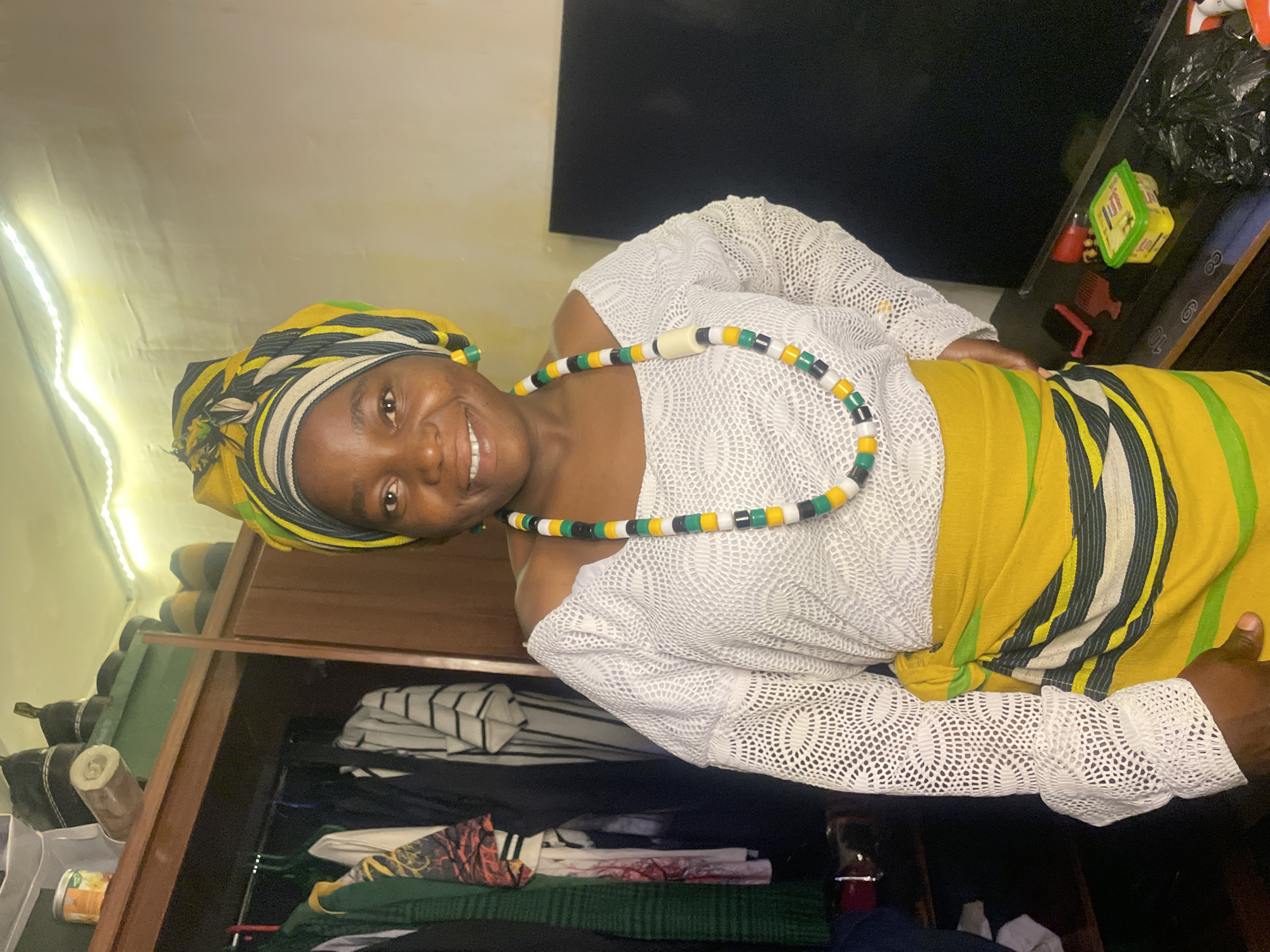 A woman with dark skin is indoors. She is smiling. She is wearing a head wrap that is yellow, green, white, and black. She has on a white lace shirt, a yellow and green sash, and a beaded necklace of the same colors. The location is unclear, but based on the clothing and skin tone, it is likely not Jos, Nigeria, where people are lighter-skinned. She is standing in front of a closet that is open and contains clothing. Next to her is a dresser or table with a black top. On the top are several objects, including a crumpled plastic bag. The lighting is bright with an unidentifiable string of lights in the background.