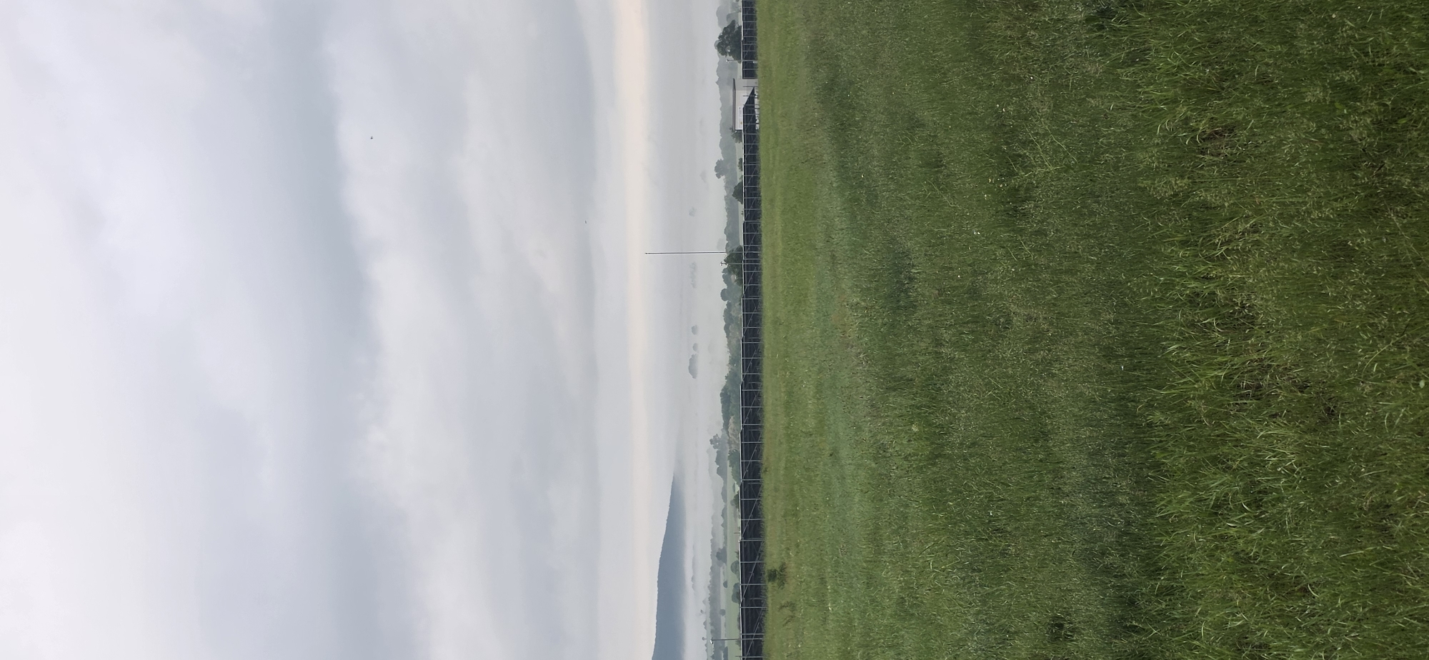 The image depicts an outdoor scene, seemingly in a rural or suburban area. The left side of the image primarily consists of a cloudy, overcast sky, with varying shades of gray. On the right side, there is a field of green grass. A fence appears to run horizontally across the middle of the image, dividing the sky from the field. In the middle distance, behind the fence, there are some trees and possibly a building. A pole rises vertically from the ground near the middle of the image. The image is rotated 90 degrees counter-clockwise. The location is Esen, Bulgaria. The scene lacks people or specific actions.