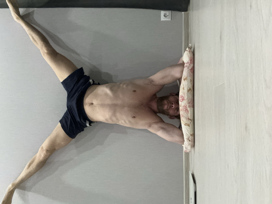 An adult male is performing a headstand against a light grey wall in an indoor setting. He is shirtless, revealing a muscular physique, and wears dark blue athletic shorts with a light stripe. His legs are spread in a wide V-shape, with his feet resting against the wall. His head is positioned downwards, supported by a small, rectangular pillow featuring a pink floral pattern on a cream background, which rests on a light-colored, wood-textured floor. His forearms are on the floor, providing additional support. To the immediate left on the floor, a white digital bathroom scale is visible. To the right, mounted on the grey wall, is a white Type F power outlet, consistent with electrical infrastructure in Saint Petersburg, Russia. A dark vertical element, possibly a doorframe, is partially visible at the far right edge of the frame.