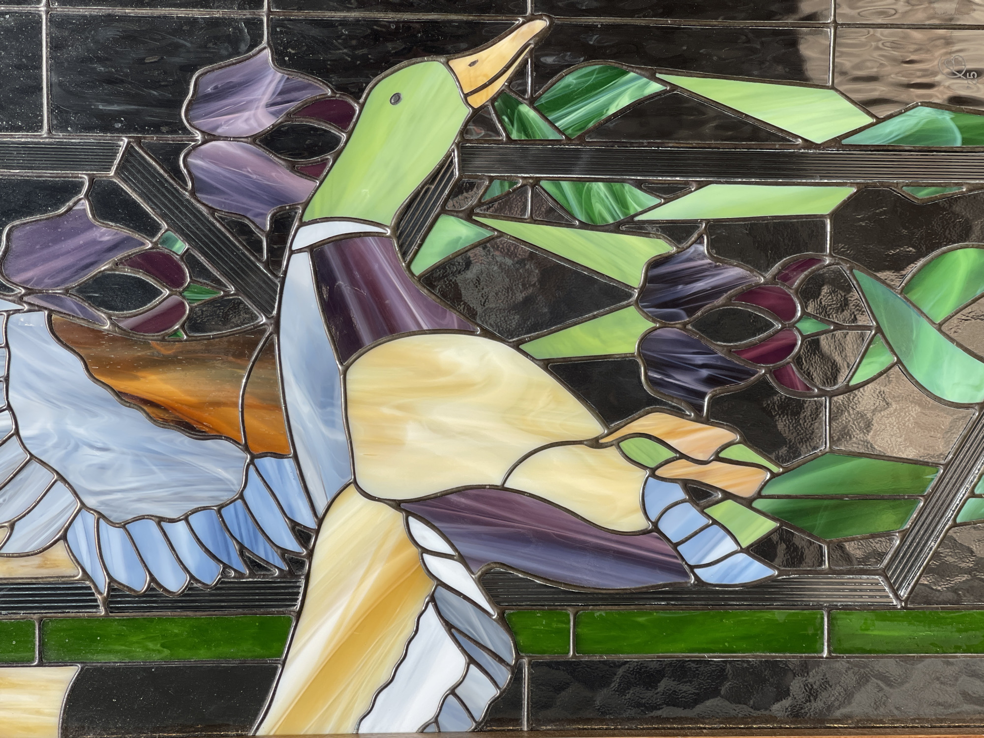 This is a close-up, eye-level shot of a stained glass panel depicting a mallard duck. The duck is rendered in profile, facing left, with its head and neck angled upwards. Its beak is a bright yellow, and its eye is a small black dot. The head and neck are a vibrant green, transitioning to a deep purple on its chest and back. The duck's body is predominantly a creamy yellow, with patches of white on its breast and undertail. Its wings are spread, showcasing a mix of pale blue and white glass pieces that mimic feathers.
The background of the stained glass is a complex arrangement of abstract shapes in various shades of green, purple, black, and orange. Some green pieces have a swirling, translucent texture, suggesting foliage or water. The purple pieces, clustered in the upper left and mid-right, resemble abstract flowers or petals. The black glass appears textured and opaque, providing a strong contrast to the lighter colors. Horizontal lines of dark glass divide sections of the panel, adding a linear element to the design.
The overall impression is a stylized and artistic representation of a duck in its natural environment, possibly a marsh or pond. The lighting appears even, highlighting the colors and textures of the glass. There is no indication of time of day or weather. The location context of Zelenogradsk, Russia, does not provide any direct visual cues within the image itself. There is a small, stylized logo with what appears to be a stylized "K" and a "5" in the upper right corner of the panel, against the black glass.