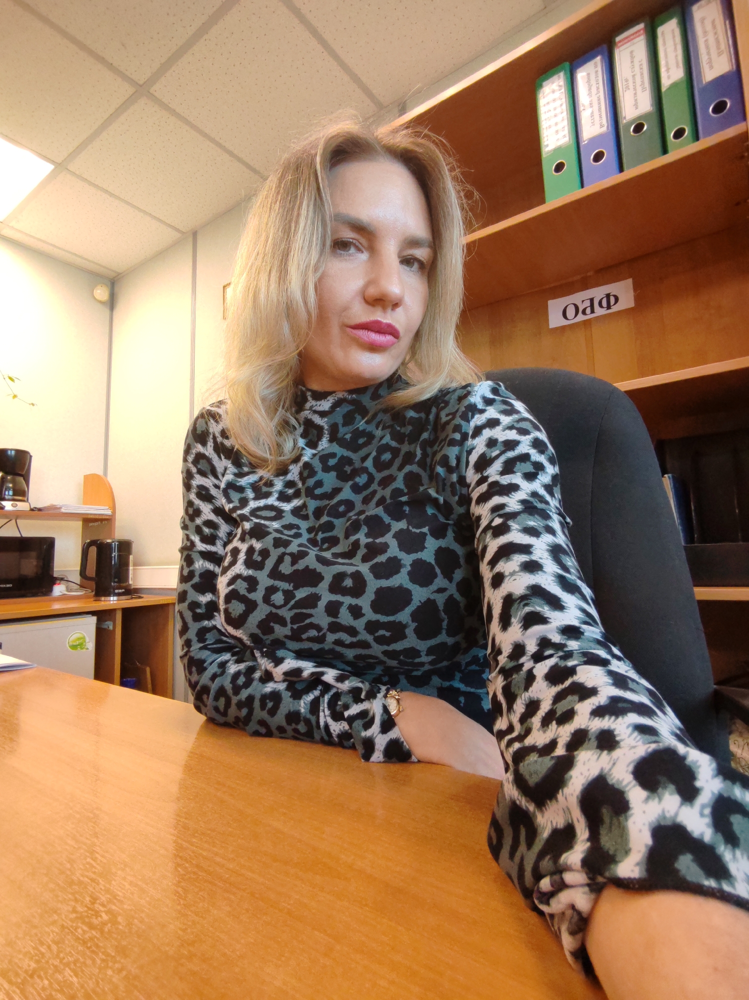 A woman with blonde hair and red lipstick is centered in the frame, taking a selfie in an indoor office setting. She wears a long-sleeved top featuring a black, grey-blue, and white leopard print. Her left arm is extended towards the camera, holding the device, while her right arm rests on a light brown wooden desk. She has a neutral to slightly serious expression, looking directly at the viewer.
The background shows a typical office environment. To her right, a wooden shelving unit displays several ring binders in green, blue, and a lighter grey/white on the top shelf. Below these, a white label with the Cyrillic letters "ОАФ" is visible. The text on the binders also appears to be in Cyrillic script.
To her left, on a lower shelf of the desk, are various appliances: a black coffee maker, a black electric kettle, and a small black electronic device that resembles a microwave. Below these, a small white refrigerator or cooler with a green label is seen. The ceiling is fitted with white, perforated acoustic tiles and fluorescent lighting. A subtle hint of green foliage from a plant is visible in the far left of the image. The time of day is not explicitly stated but suggests interior daytime lighting.
