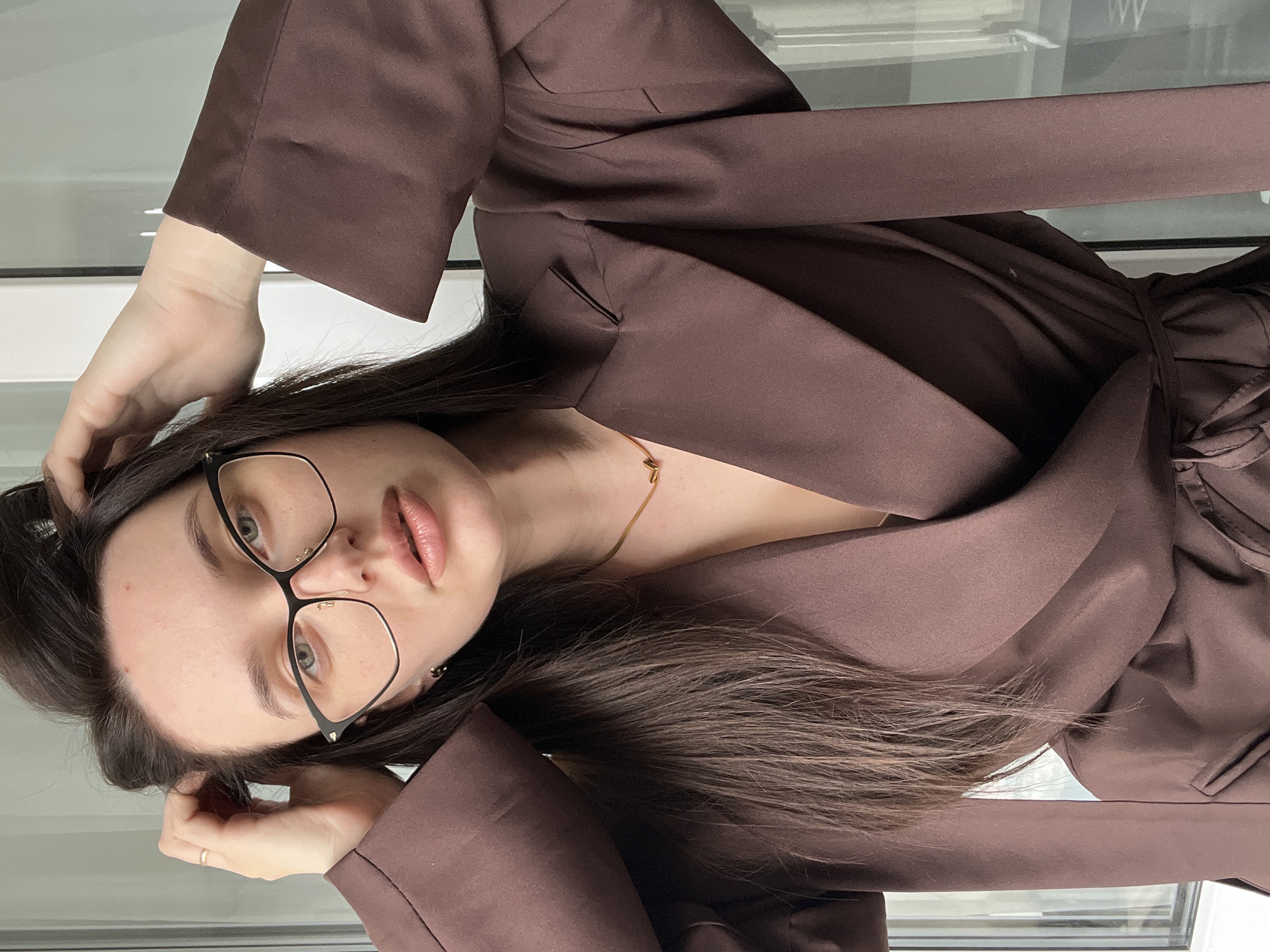 A young woman with long, dark hair and wearing glasses is posing indoors. She is dressed in a brown wrap dress or blazer-style outfit. The setting appears to be near a window or glass panel, likely indoors. The location appears to be Tyumen, Russia.