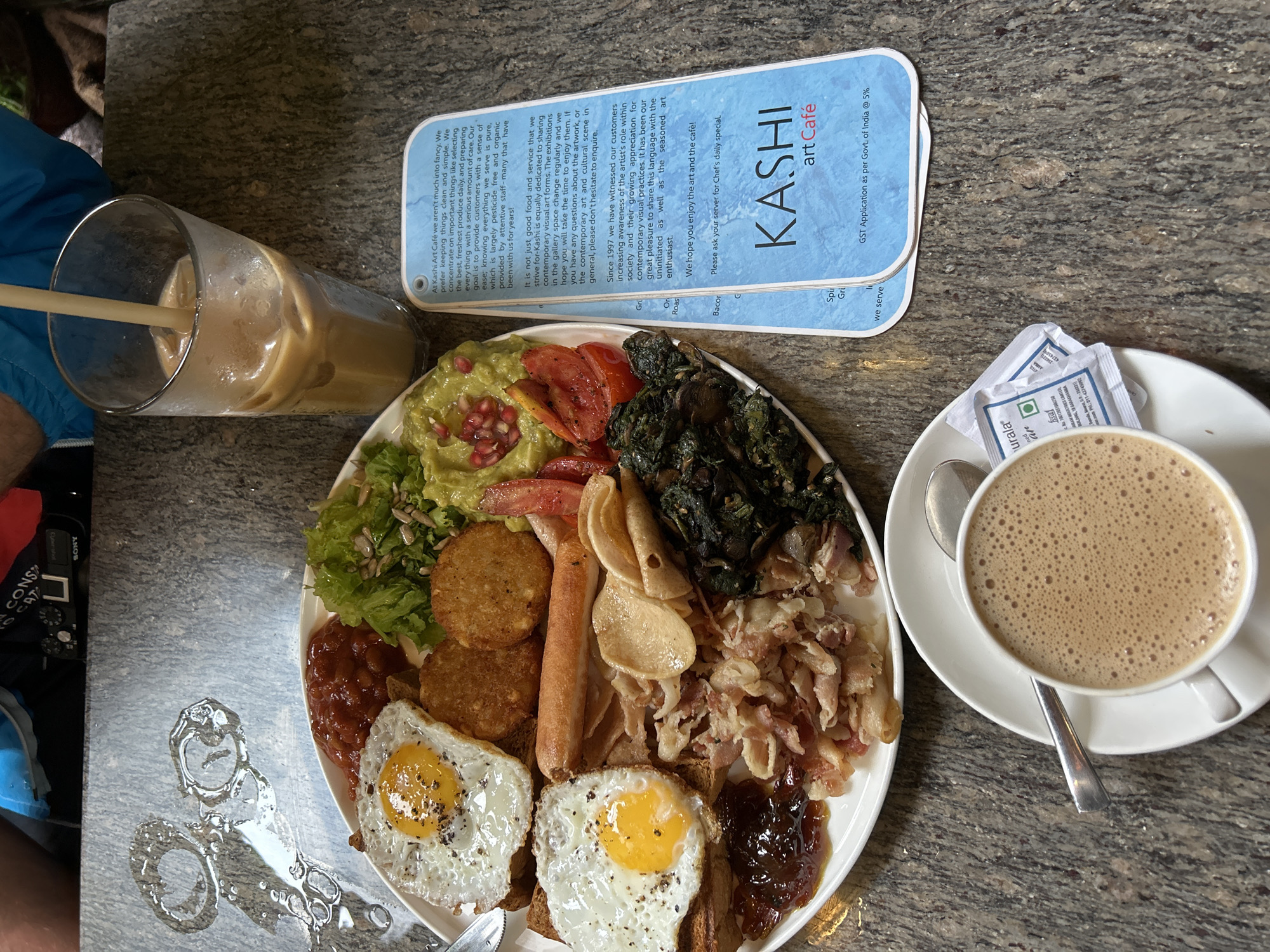 An overhead shot captures a rich breakfast or brunch spread at Kashi Art Café in Kochi, India. The main subject is a large white oval platter laden with a diverse array of food. On the platter, there are two perfectly fried eggs, two golden-brown hash browns, a sausage, crumbled bacon bits, baked beans, and a dollop of jam or preserve. Complementing these are fresh components including guacamole topped with pomegranate seeds, a mixed green salad with sunflower seeds, sliced tomatoes, and sautéed dark green leafy vegetables, along with some thin, cracker-like flatbread pieces.
To the left of the platter is a tall glass of iced coffee with a straw, partially obscured by a person's blue jacket. On the right, a white ceramic cup of frothy hot coffee sits on a matching saucer, accompanied by a spoon and two "naturala" branded sugar packets.
Above the food and drinks, a light blue information card for "KASHI art Café" is displayed. The card describes the cafe's commitment to using "freshest produce daily," "pesticide free and organic" ingredients, and its role in "sharing contemporary visual art forms" through changing exhibitions since 1997. It also notes "GST Application as per Govt. of India @ 5%." The dining surface is a dark, speckled granite or stone tabletop. A notable detail on the table is a water stain shaped like scissors to the left of the food platter. A "SONY" branded camera is also partially visible in the upper left background. The overall scene suggests a relaxed and hearty meal.