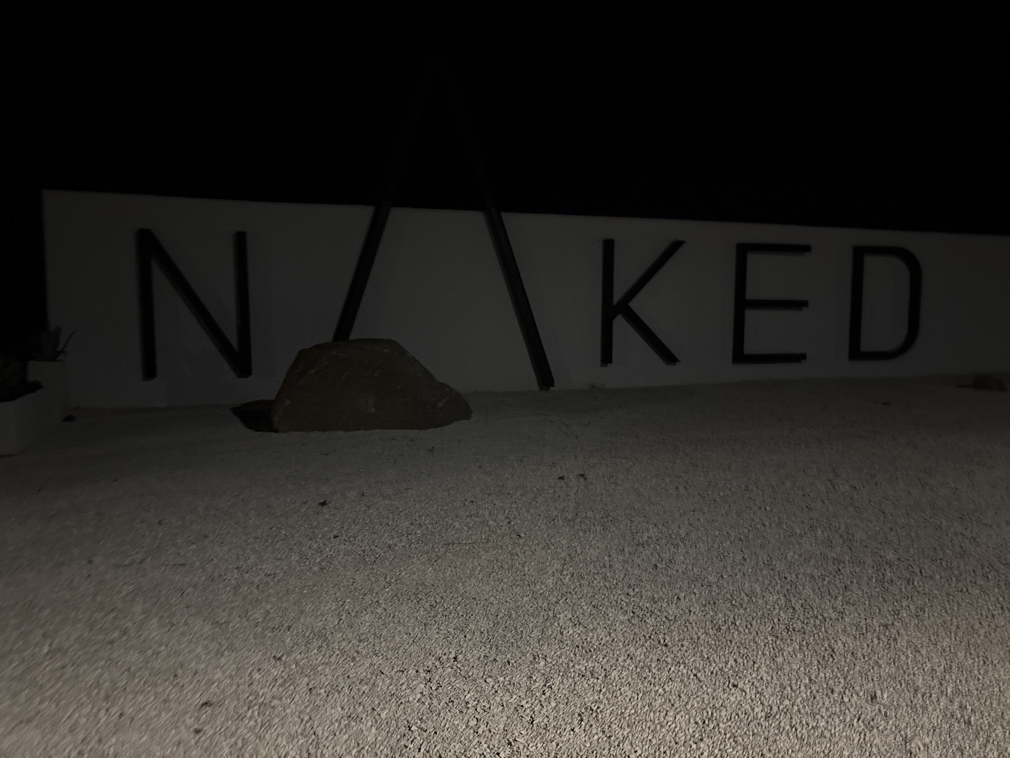 The image shows a sign that reads "NAKED". The sign is mounted on a white background and the letters are black. There is a rock in front of the sign, and the ground is covered in small pebbles. The image is taken at night, and the lighting is dim. There is no information in the image to suggest it is located in Burgas, Bulgaria.