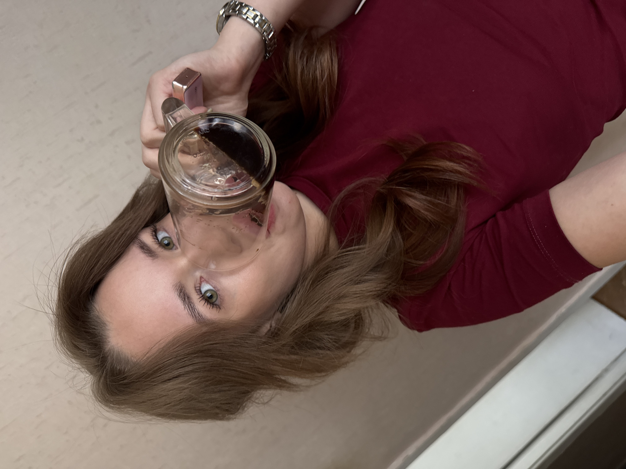 A selfie taken by a young woman with shoulder-length brown hair and green eyes, wearing a dark red t-shirt. She is holding a glass mug filled with a dark liquid, possibly coffee or tea, and drinking from it. Her left hand, adorned with a silver watch, holds the mug. The background appears to be an interior wall with a textured, light beige wallpaper. The lighting is soft, suggesting an indoor setting. The image does not provide any specific visual cues that would allow for the identification of the location in Chaikovsky, Russia.