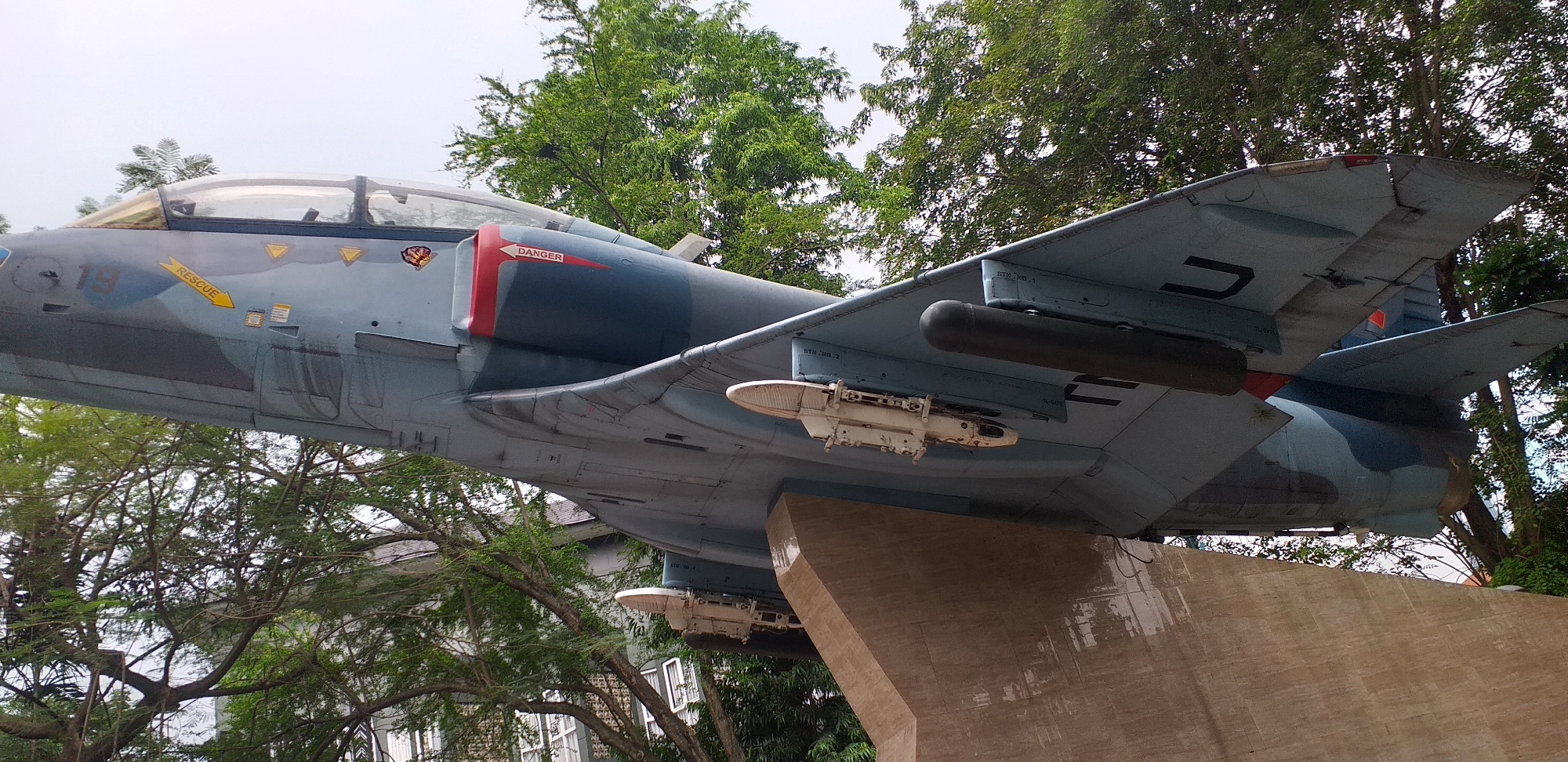 A Douglas A-4 Skyhawk, painted in a two-tone blue camouflage pattern, is mounted on a concrete plinth. The aircraft features a "RESCUE" label with an arrow pointing towards the ejection seat area, a red "DANGER" marking around the air intake, and hardpoints for ordnance under the wings. The aircraft is located in South Jakarta, Indonesia. The plinth is damp and has a visible texture. Trees surround the monument, obscuring the sky. A building is partially visible through the trees to the left of the image.