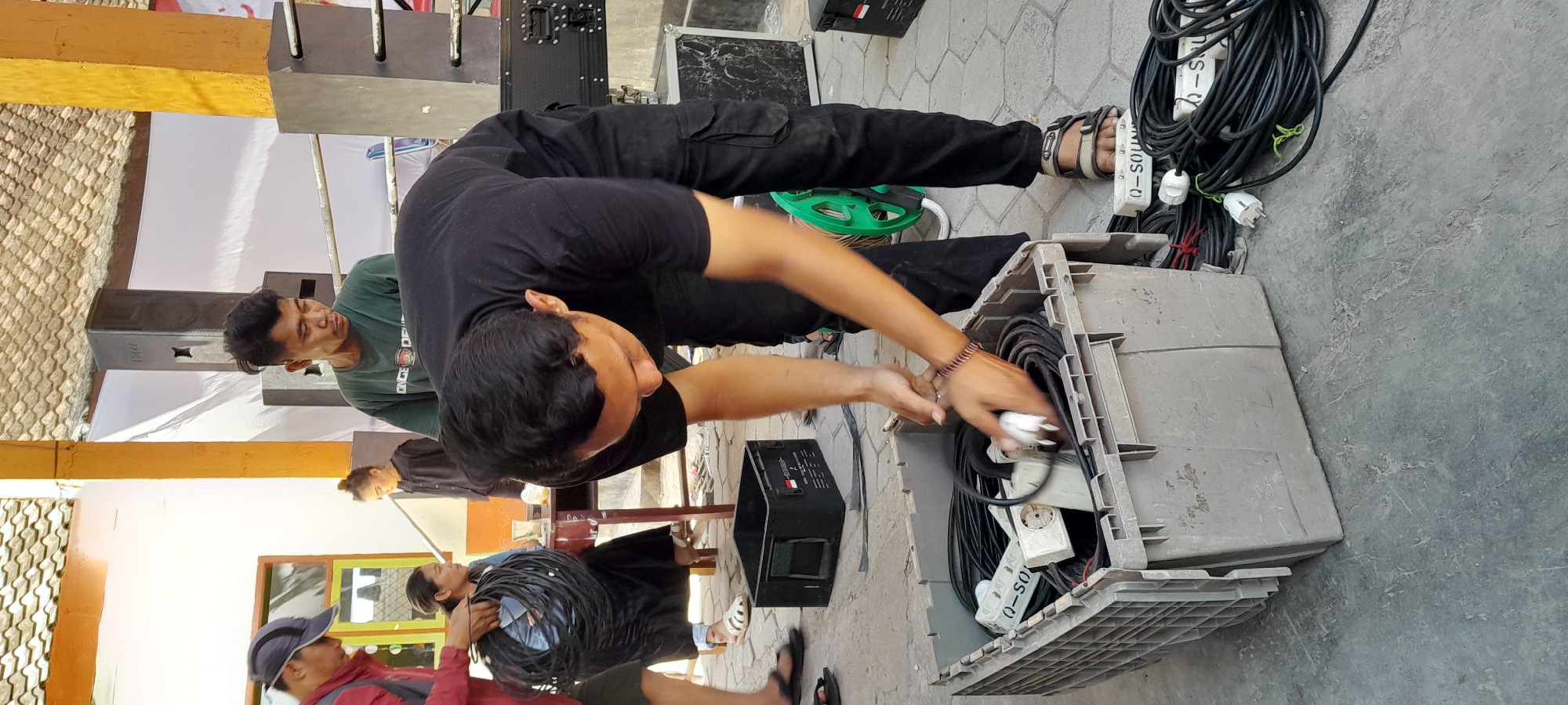 In Lemahbang Dewo, Indonesia, a man in a black t-shirt and cargo pants is bent over a gray plastic crate filled with coiled black electrical cables and white power strip extenders. He is holding a white plug in his right hand, appearing to be connecting or disconnecting it from a cable. The crate is on a paved ground surface, surrounded by more coiled cables, some of which have "Q-SOUND" printed on white rectangular casings. To the left, another man wearing a red shirt and a blue cap is holding a large coil of black cable. In the background, other individuals are visible, along with what appear to be audio speakers and a table. The scene is outdoors, with a tiled roof overhead and part of a building on the left.