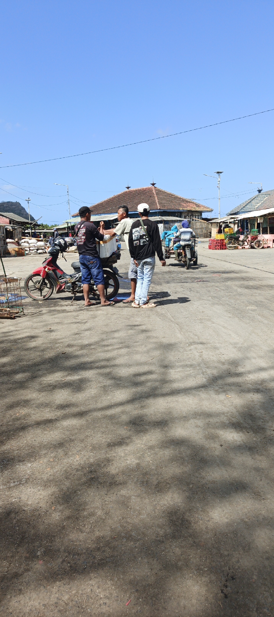 In Puger, Indonesia, under a clear blue sky, a busy marketplace scene unfolds. Three men stand around a red motorcycle, one holding a white cooler, and another man wearing a black shirt and a white cap. The marketplace appears to be a hub of activity, with buildings and stalls visible in the background. A man is riding a three-wheeled vehicle, and various crates and goods are scattered around the area. The ground is paved, with shadows from overhead trees or structures cast across it.