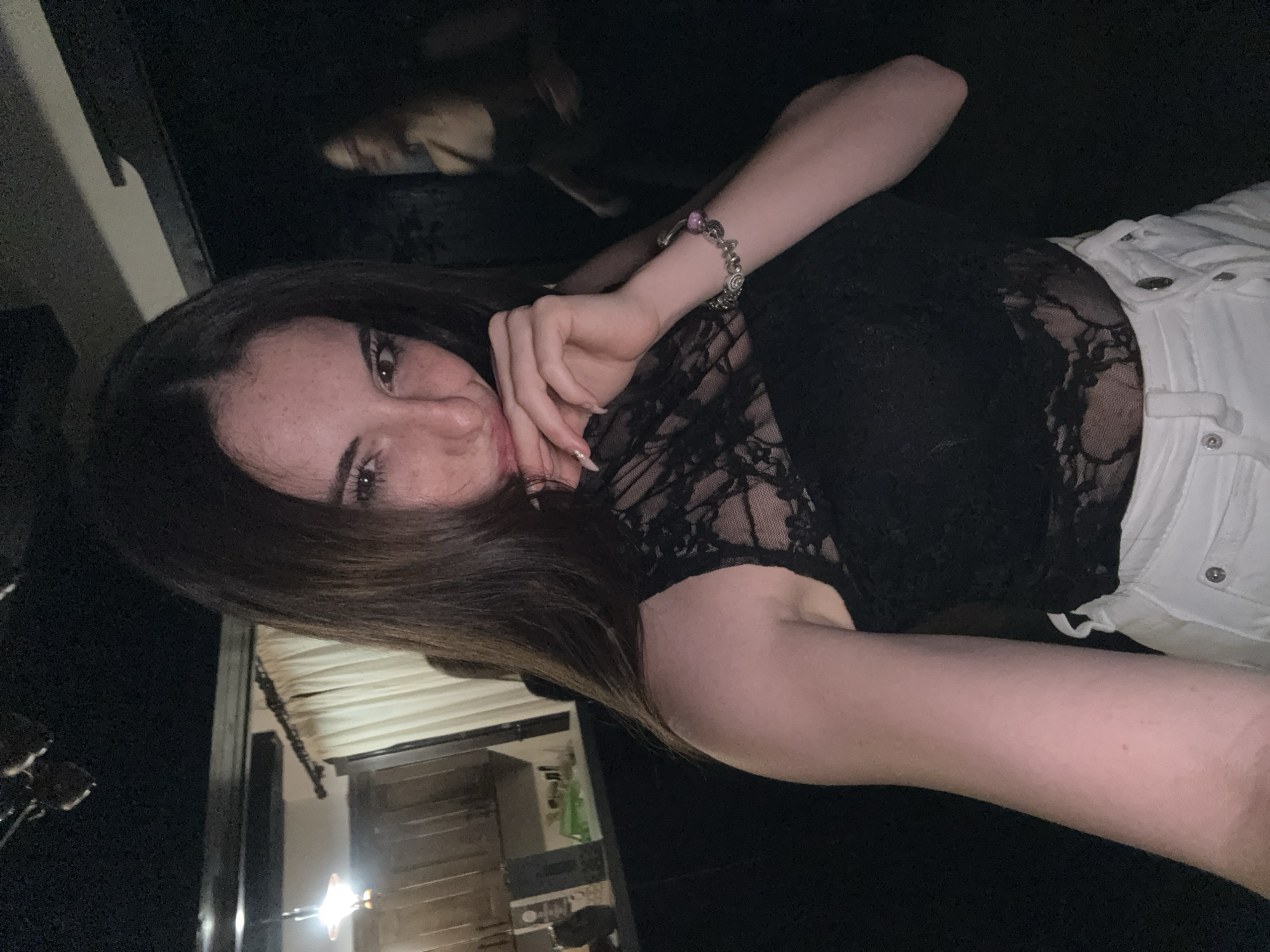 The image depicts a young woman posing for a selfie indoors, likely at a bar or nightclub. She has long, dark hair and is wearing a black lace top and white shorts. Her right hand is near her mouth, and she is wearing a bracelet on her left wrist. In the background, another person is visible, blurred and out of focus, and the environment appears to be dimly lit with some wooden elements and light fixtures. The location is identified as Tazha, Bulgaria.