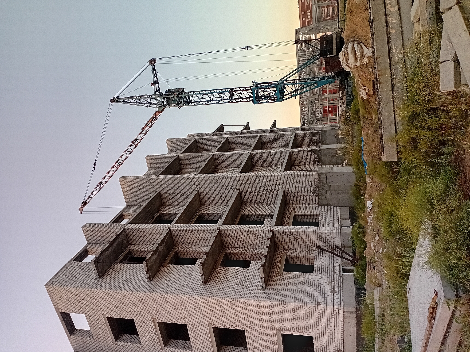An outdoor, low-angle, vertical shot captures the construction of a multi-story building, likely an apartment complex, in Semey, Kazakhstan. A prominent turquoise and red tower crane dominates the right side of the frame, its boom extending over the unfinished structure. The building itself is constructed of light-colored bricks with dark, open window frames and concrete balconies. Piles of dirt, sandbags, and concrete slabs are visible on the ground, along with overgrown green grass and weeds, suggesting the construction site is in an undeveloped area. In the background, other partially constructed buildings and skeletal steel frameworks are discernible under a pale, clear sky. The lighting suggests it is either early morning or late evening. There are no people visible in the scene.