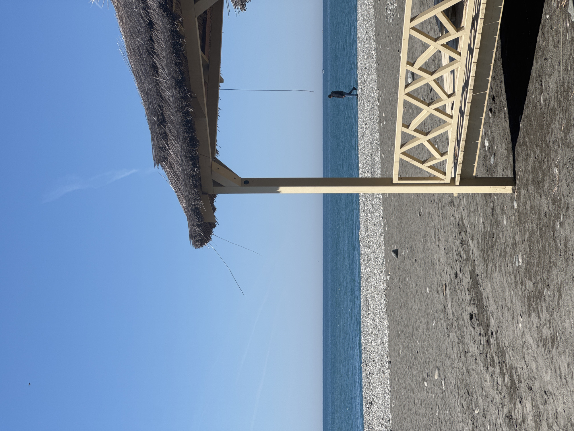 The image captures a bright, sunny beach scene, oriented with the horizon line running horizontally across the middle. The upper half of the frame is dominated by a clear, vibrant blue sky, featuring a faint contrail in the distance.
Below the sky, the calm, blue waters of the sea extend across the left side of the image, meeting a dark pebble and sand beach on the right. A solitary figure is visible in the mid-ground, walking along the shallow water's edge, facing away from the viewer.
In the upper left corner, the edge of a thatched roof extends into the frame, suggesting a beach hut or umbrella. A thin wire or string hangs down from this thatched structure. On the right side, a light-colored wooden structure with a distinctive lattice pattern is prominent, casting a sharp shadow onto the dark sand. This structure appears to be a railing or part of a walkway on the beach. The beach surface consists of dark, textured sand interspersed with numerous pebbles.
The scene depicts a peaceful, warm day at the seaside, likely midday or early afternoon given the bright sunlight and clear shadows. There is no visible text in the image.