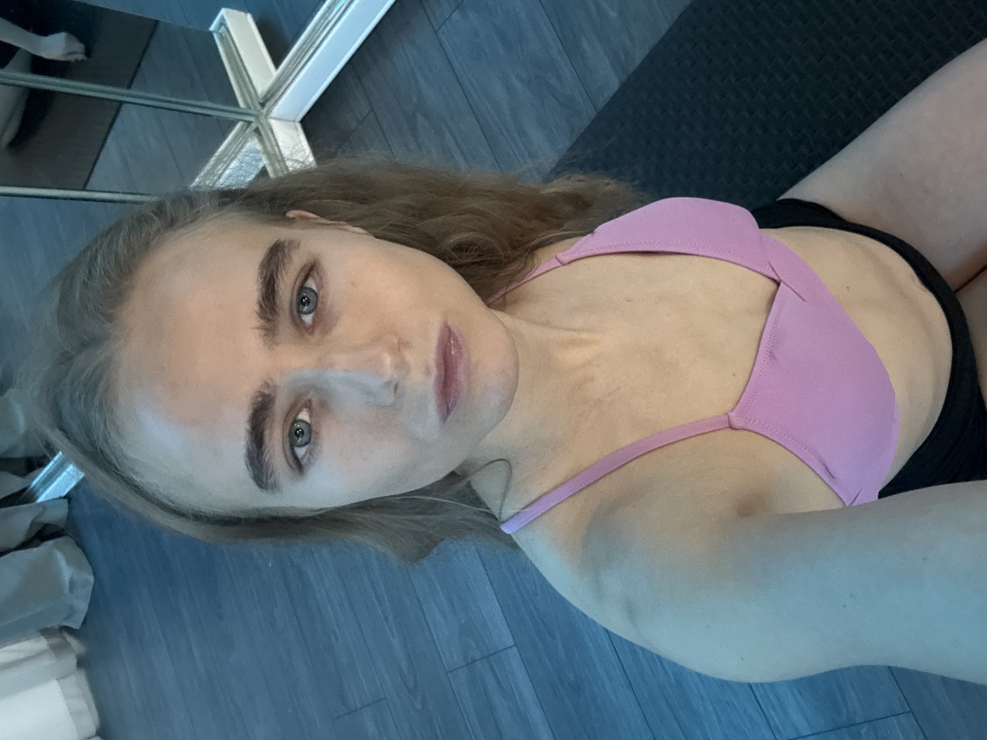 A young fair-skinned person with shoulder-length light brown hair and blue eyes is facing the camera. They are wearing a pink sports bra. A black mat is visible in the background. The location is Unknown, Unknown.