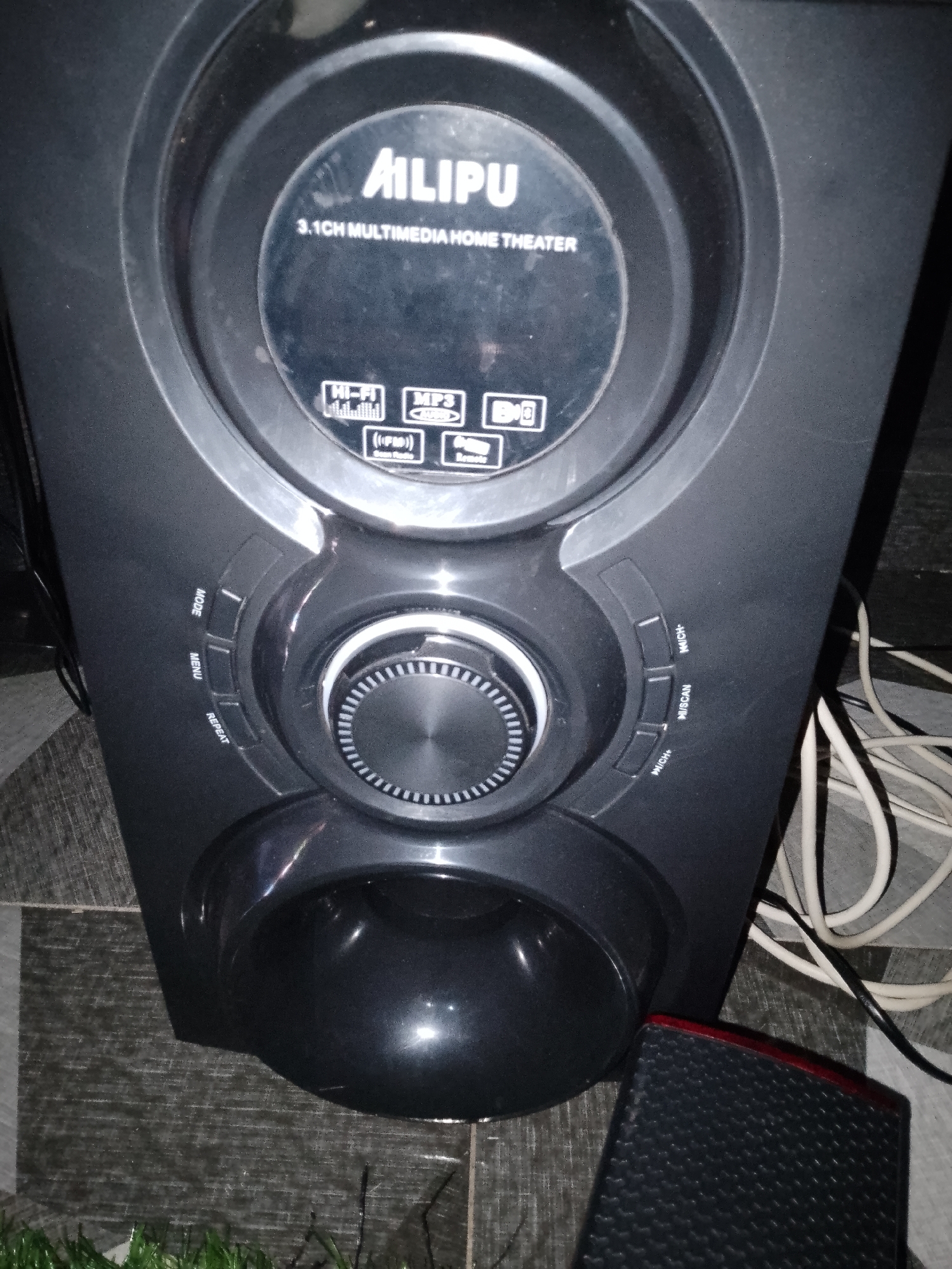 An AILIPU 3.1CH MULTIMEDIA HOME THEATER speaker is captured in a close-up, low-angle shot. The speaker is black with a circular display in the center, which features various icons indicating Hi-Fi, MP3, FM Radio, and Remote capabilities. Below the display, a large volume knob is prominently positioned, surrounded by several control buttons labeled "MODE," "MENU," "REPEAT," "M/CH+," "M/SCAN," and "M/CH-." A glossy, oval-shaped bass reflex port is located at the bottom of the speaker. To the right of the speaker, white and black power cords are visible, lying on a tiled floor with a grey and white checkered pattern. Part of a red-trimmed black speaker is visible in the lower right corner of the image. The background is dark and out of focus. The scene is indoors, with no discernible urban context or city.