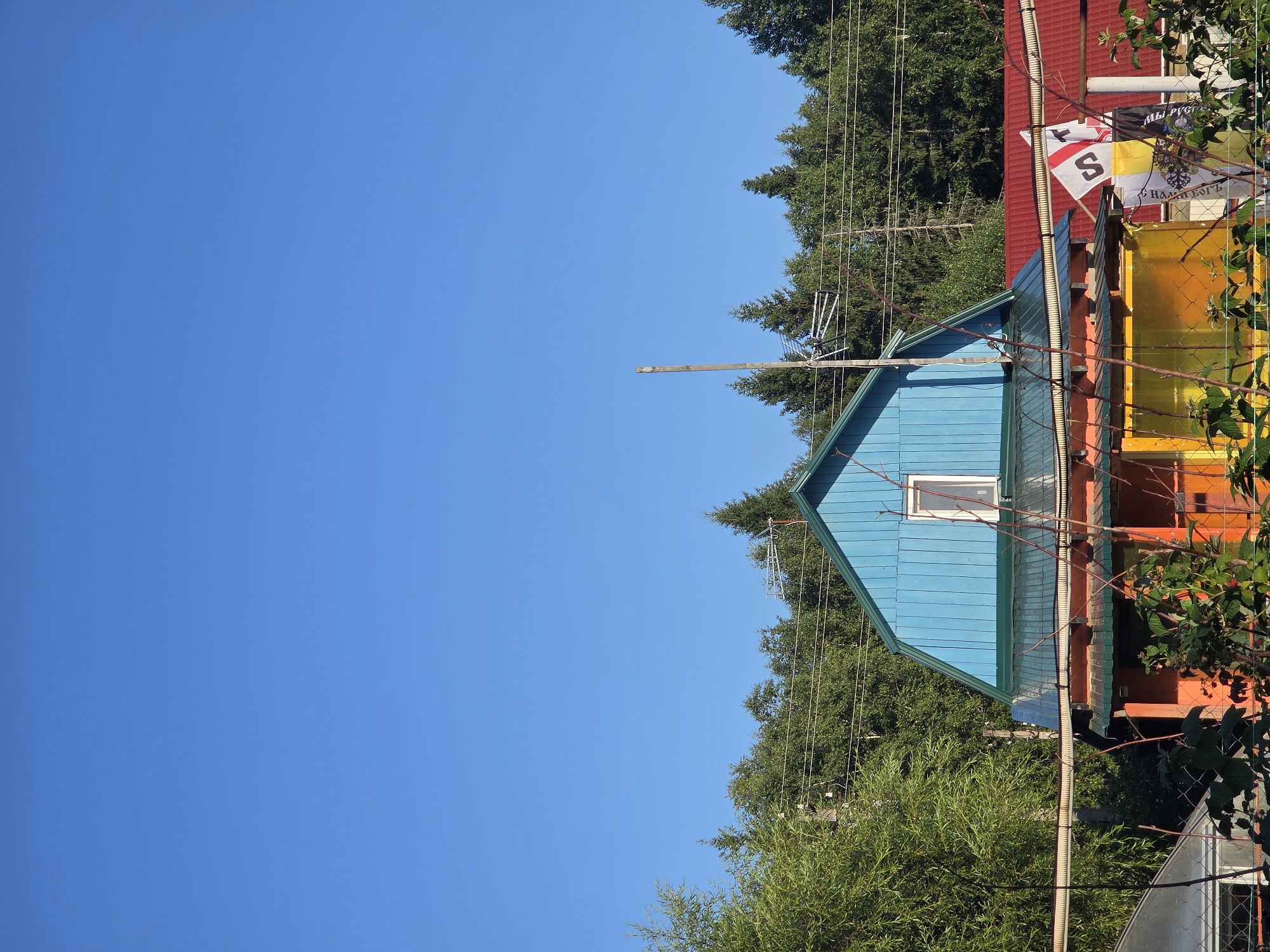 A bright, clear blue sky dominates the upper portion of the image. Below, a two-story house with a blue, gabled roof and a green metal roof on the lower level is visible. A single window is present on the upper blue section of the house. The lower part of the house is painted orange and yellow, with a metal roof that appears to be red. Several flags are attached to poles in front of the house; one is white with red markings, including a prominent "S". Another flag is yellow and black with Cyrillic lettering. A wooden pole with an antenna and wires runs vertically in front of the house, extending into the sky. Trees and foliage surround the house, with power lines strung between poles in the background. A chain-link fence and some thorny branches are in the foreground, partially obscuring the lower part of the scene. The image appears to be taken on a sunny day in Frolovo, Russia.