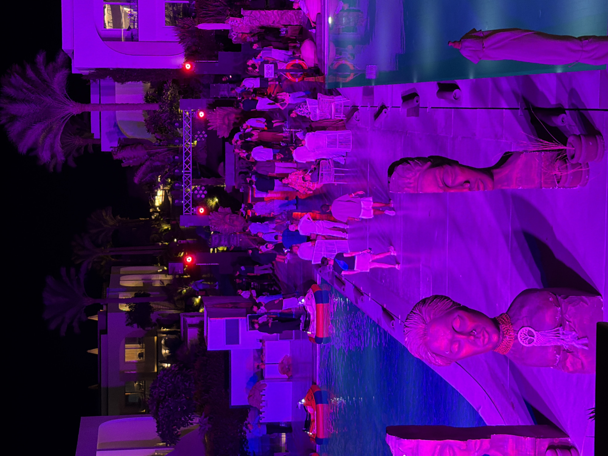 The image captures a vibrant nighttime event at a resort in Sharm El Sheikh, Egypt. The scene is dominated by purple lighting, creating a festive atmosphere. A swimming pool is visible on the left side of the image, with two red and yellow bumper boats floating in it. Several people are gathered around the pool area, some standing and others sitting on white chairs.
A walkway, also illuminated in purple, runs alongside the pool. On this walkway, a group of people are socializing and walking. Two large, sculpted busts are prominently displayed on the left side of the walkway, adding an artistic element to the scene. In the background, buildings typical of a resort in Sharm El Sheikh are visible, with some windows lit from within. Palm trees, characteristic of the region, are also present, their silhouettes dark against the night sky. A stage with lighting equipment suggests a performance or DJ set might be part of the event. The overall impression is one of a lively outdoor party or gathering in a tropical setting.