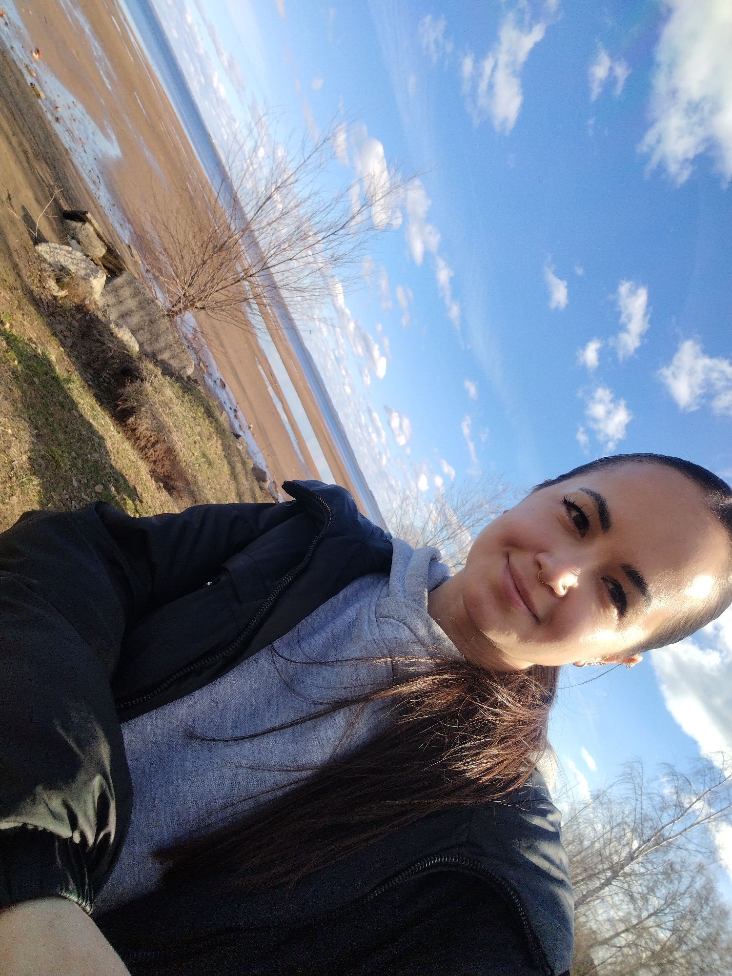 A selfie-style image shows a woman in a black jacket over a grey hoodie, looking at the camera with a slight smile. The background reveals a landscape with a body of water, possibly a river or a lake, with a sandy or muddy shore. The sky is blue with scattered clouds. A bare tree and vegetation are visible on the bank. The scene is Chaikovsky, Russia.