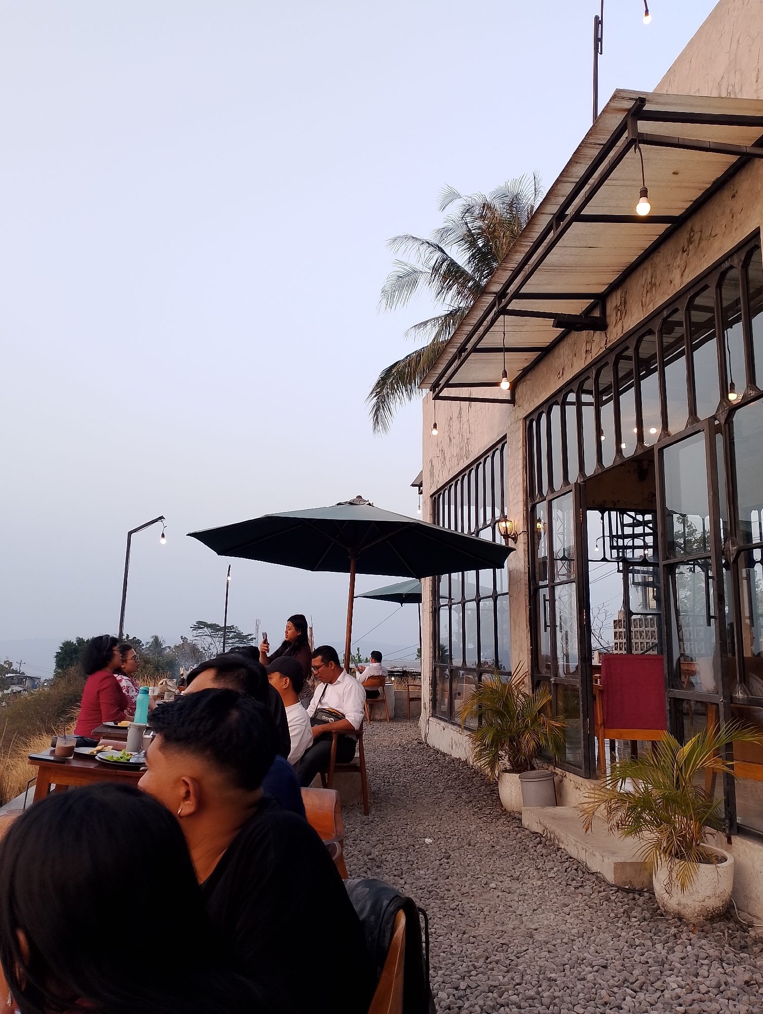 The image depicts an outdoor seating area of a cafe with a view of the city of Srimulyo, Indonesia. The cafe features large glass windows, an awning with exposed bulbs, and a gravel-covered ground. Several groups of people are seated at tables, some engaged in conversation, others looking at their phones, and one woman is taking a photo. One table has plates, drinks, and a water bottle. Two large umbrellas provide shade over some of the tables. In the background, the landscape of Srimulyo is visible with buildings and greenery. A palm tree is also present in the scene. The sky is overcast, suggesting it might be late afternoon or early evening.